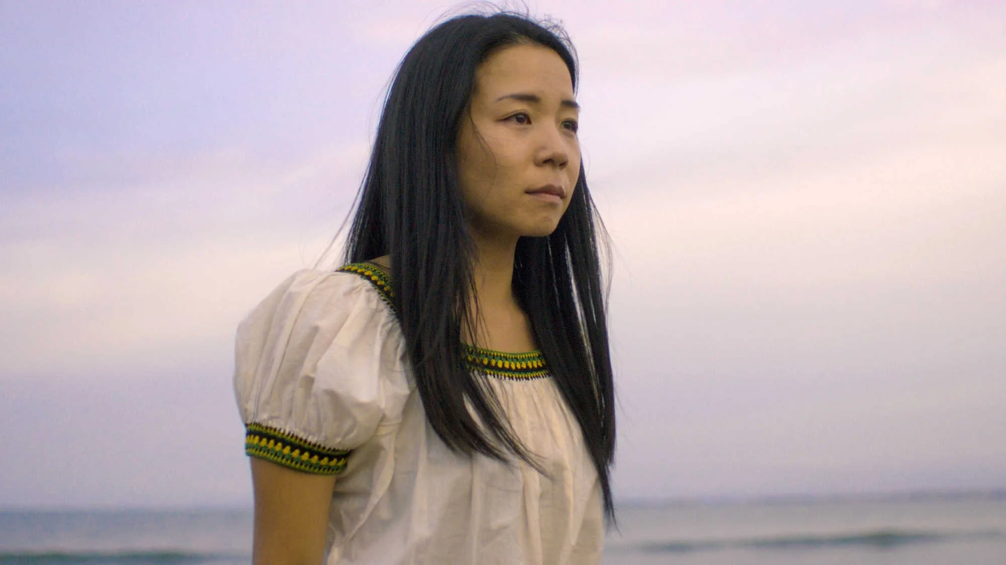 An asian woman with long black hair wearing a cream-colored dress with embroidered decorative trim, standing outdoors against a background of a cloudy sky and ocean.
