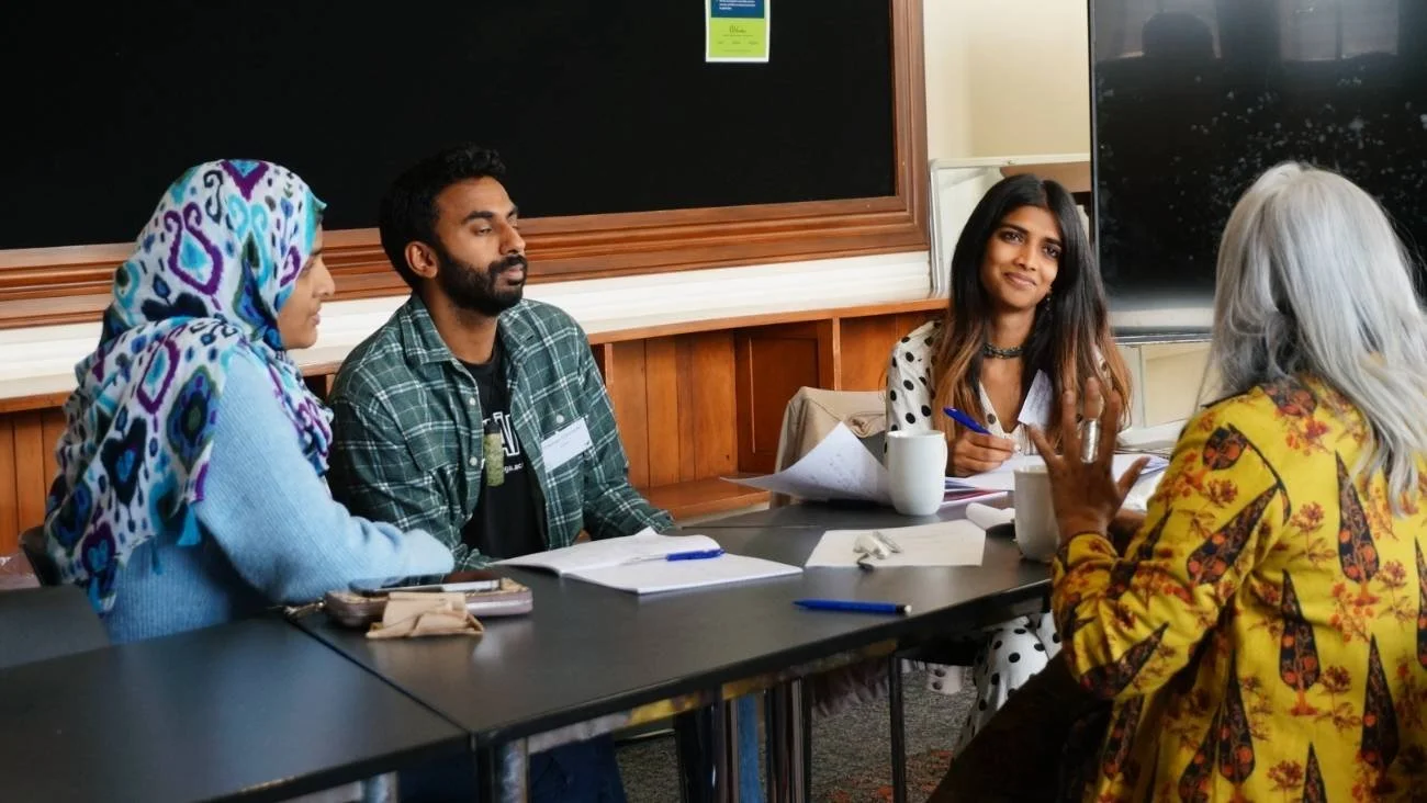 Three female Short Cuts Development Lab participants and Shuchi Kothari sit at a conference table, with notebooks and pens in front of them, in a room with a large blackboard and a marker board.