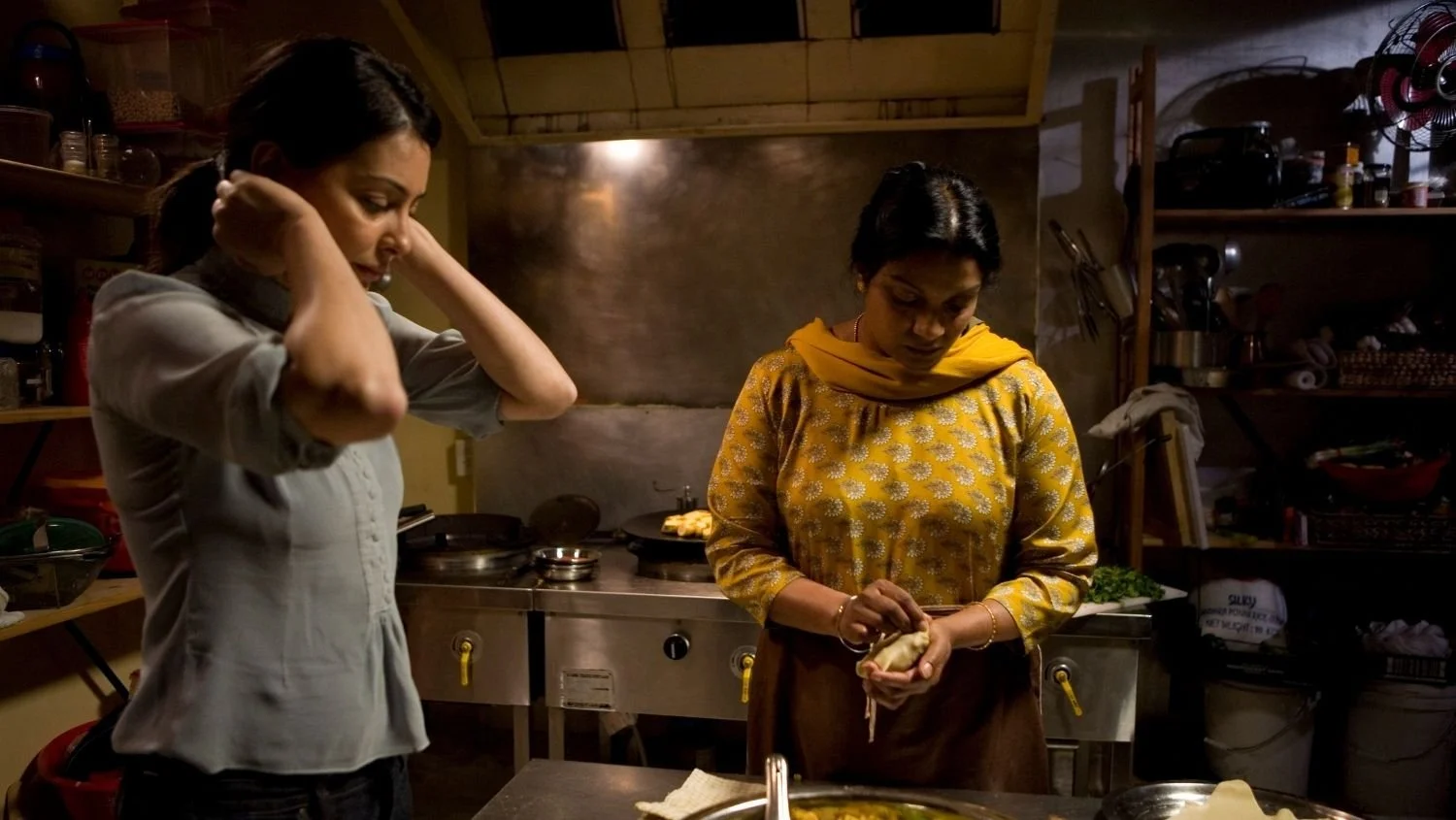 Two women preparing food in a kitchen, with shelves of utensils and ingredients around them.