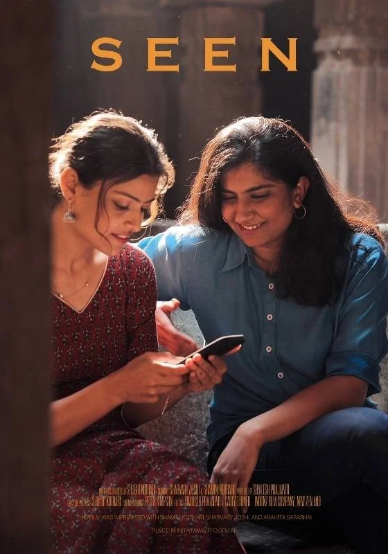 Two women sitting close together looking at a smartphone, with the word 'SEEN' above them in large letters.