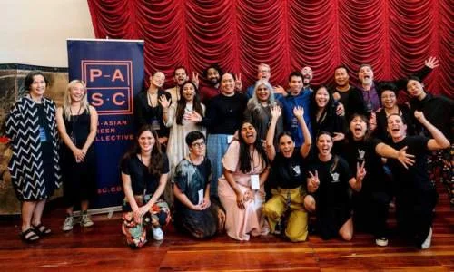 Participants in "Episode One" a year-long web-series development and pilot production programme run by Shuchi Kothari for PASC smile and wave in front of a red curtain, standing next to a PASC pull-up banner.