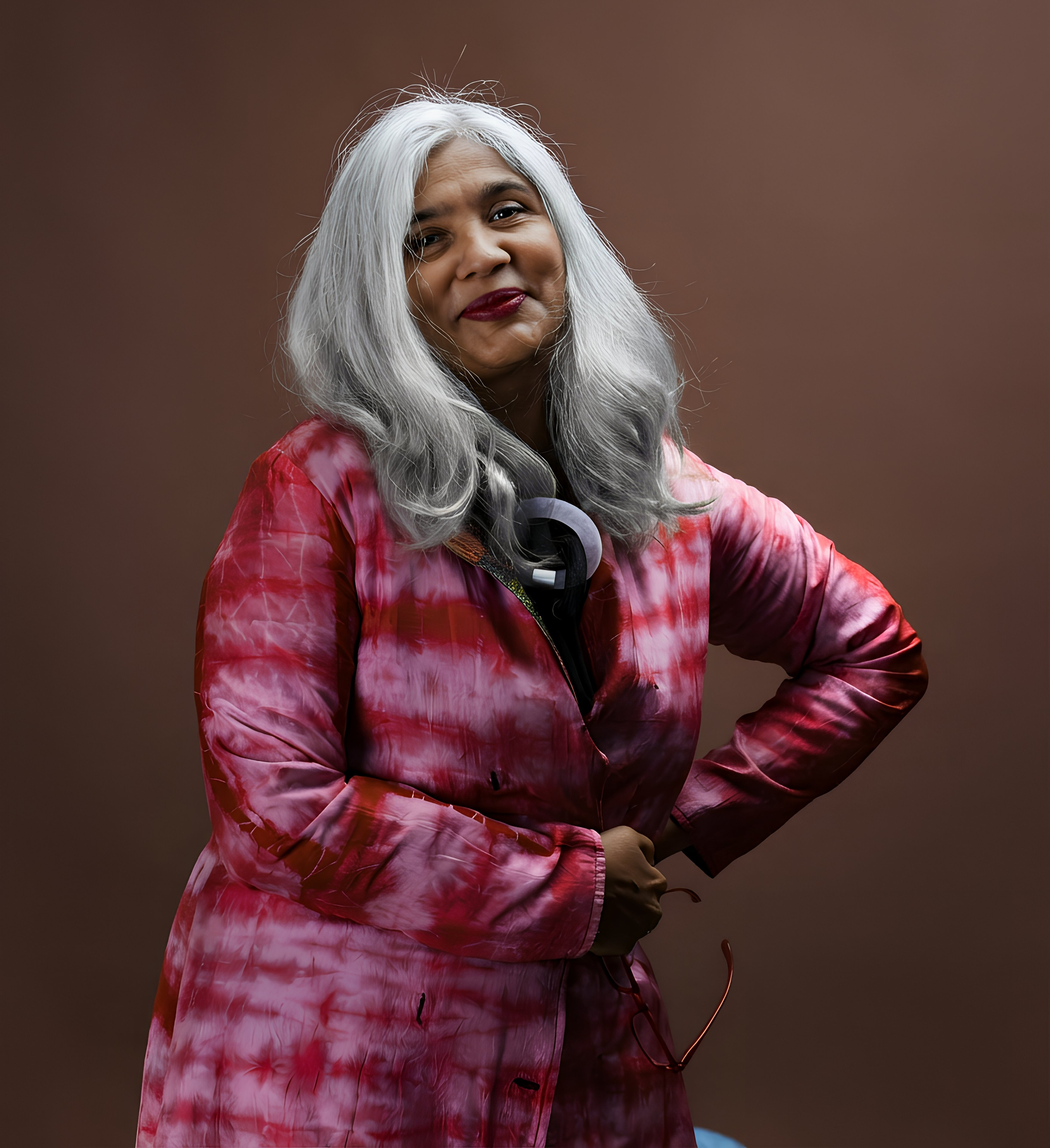Shuchi Kothari, wearing a tie-dye pink and purple jacket, standing against a brown background, smiling.