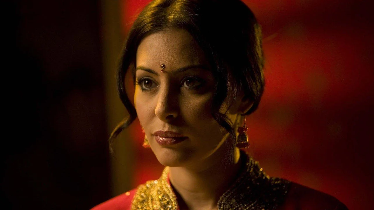 A woman in traditional Indian attire with a bindi on her forehead, wearing earrings and a richly decorated sari, in a dimly lit setting with warm red and yellow lighting.