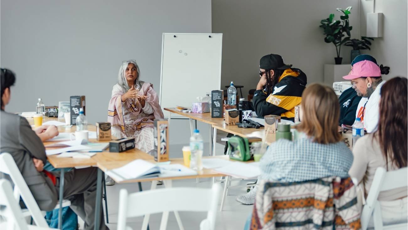 Shuchi Kothari speaks to participants at Ngā Aho Shorts Development Lab, in a modern office setting with a whiteboard and indoor plants.