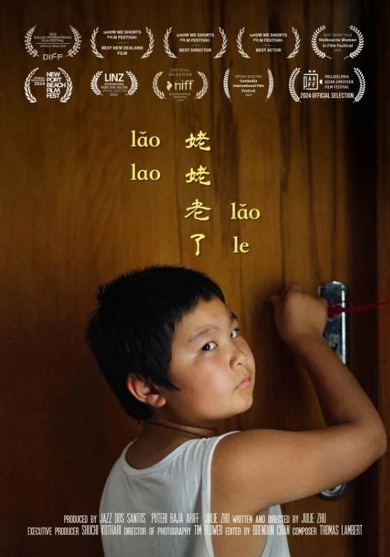 Poster for the short film Lǎo-Lao-Lǎo-Le.A young boy looking over his shoulder at the camera while drawing Chinese characters with a red marker on a wooden door. The door has the Pinyin word 'lǎo lǎo lǎo le' written alongside the Chinese characters.