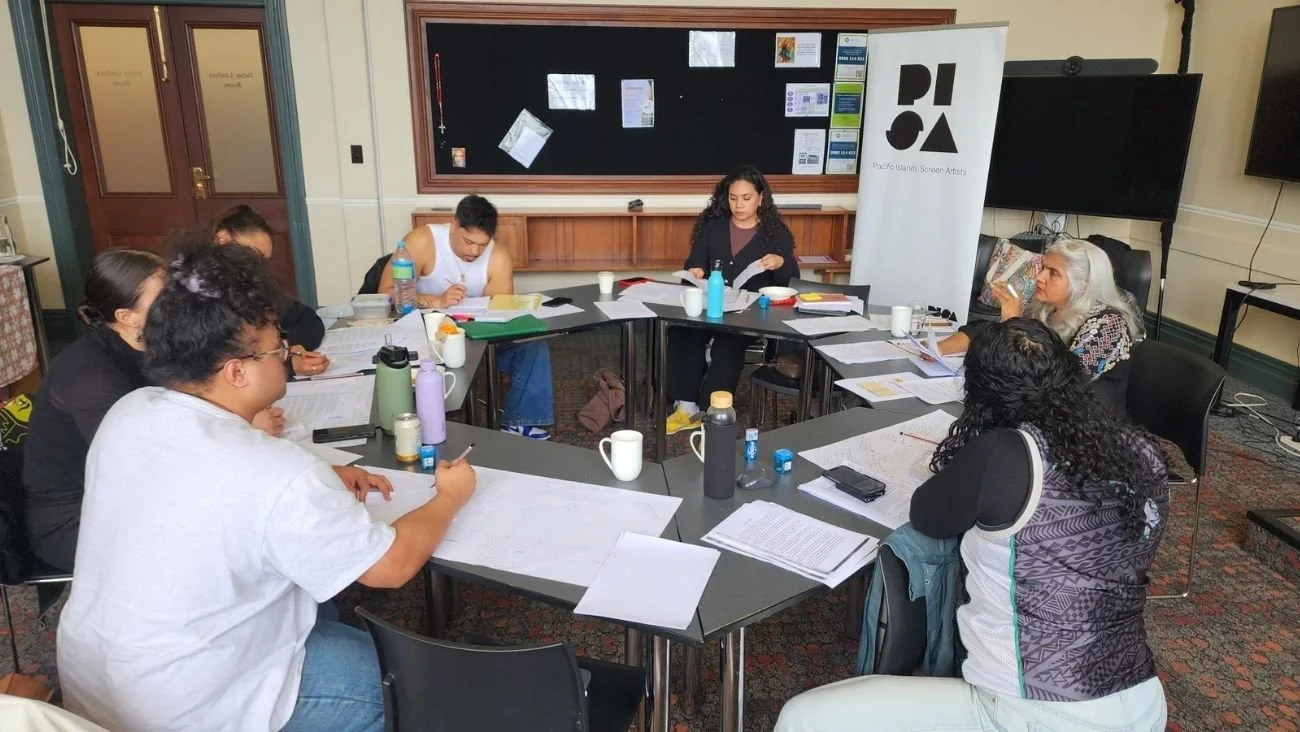 Niu Waves: Feature Film (PISA – Pacific Islanders in Screen Arts, 2025) participants listen to Shuchi Kothari present. They are engaged with papers, notebooks, and water bottles, in a room with a blackboard and a standing banner that says 'PISA'.