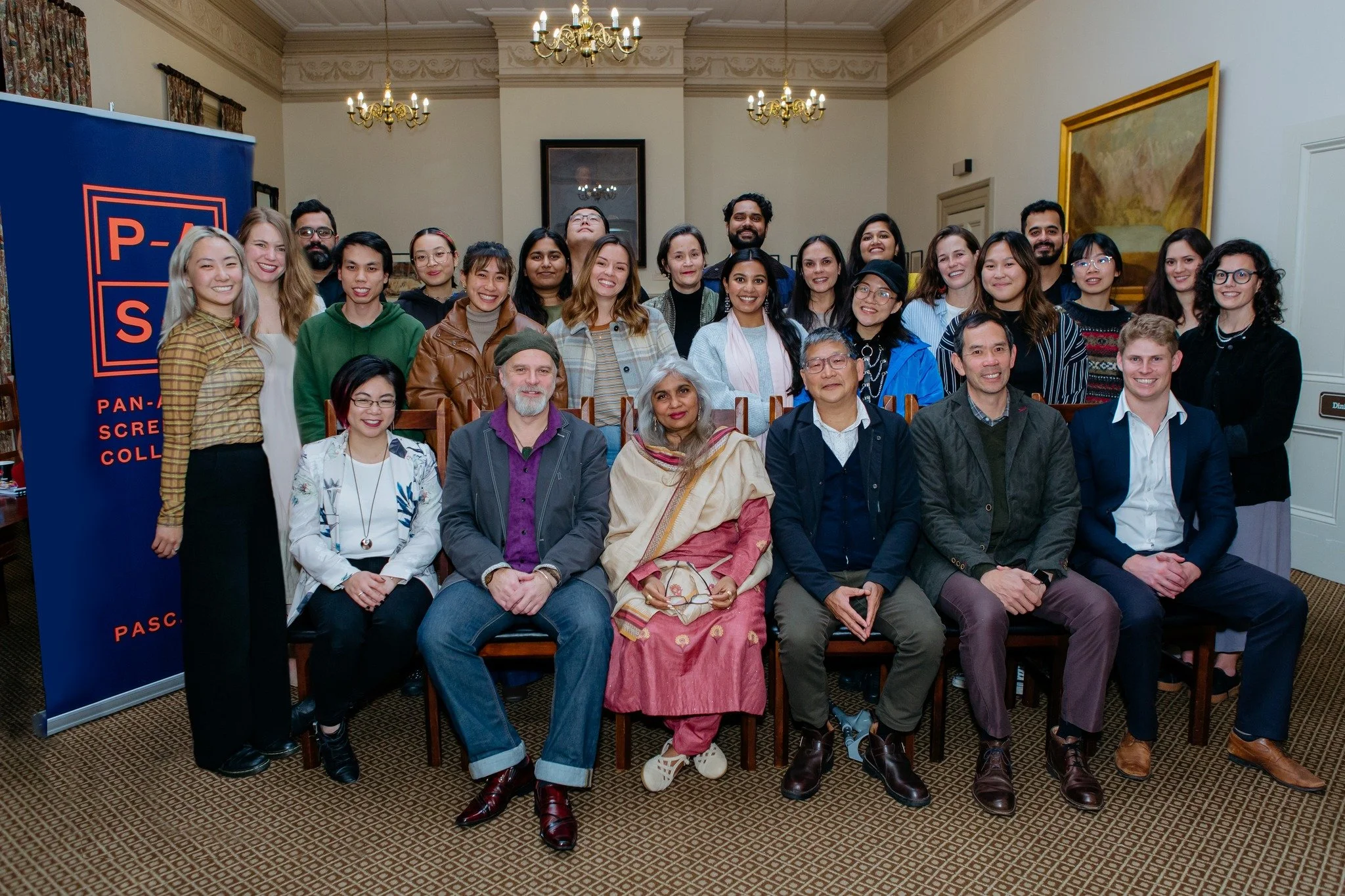 A group of about 25 PASC members pose in a formal room with chandeliers and paintings.  Shuchi Kothari sits in the center of the front row. A PASC banner is partially visible on the left side of the image.