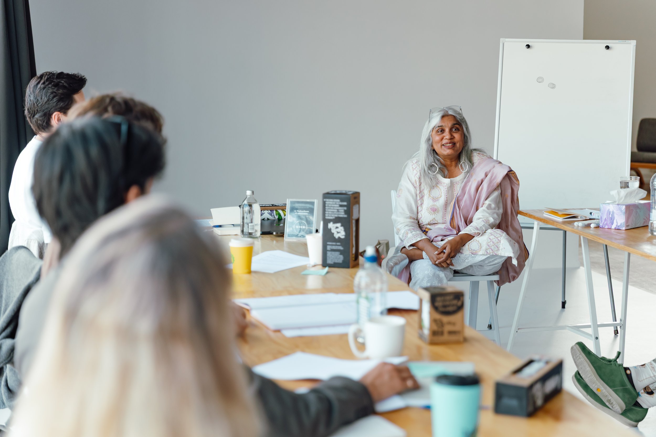 Shuchi Kothari, wearing a light-colored sari, sits at a table during a workshop, with several participants sitting in the foregroundwith a whiteboard and various items on the table.