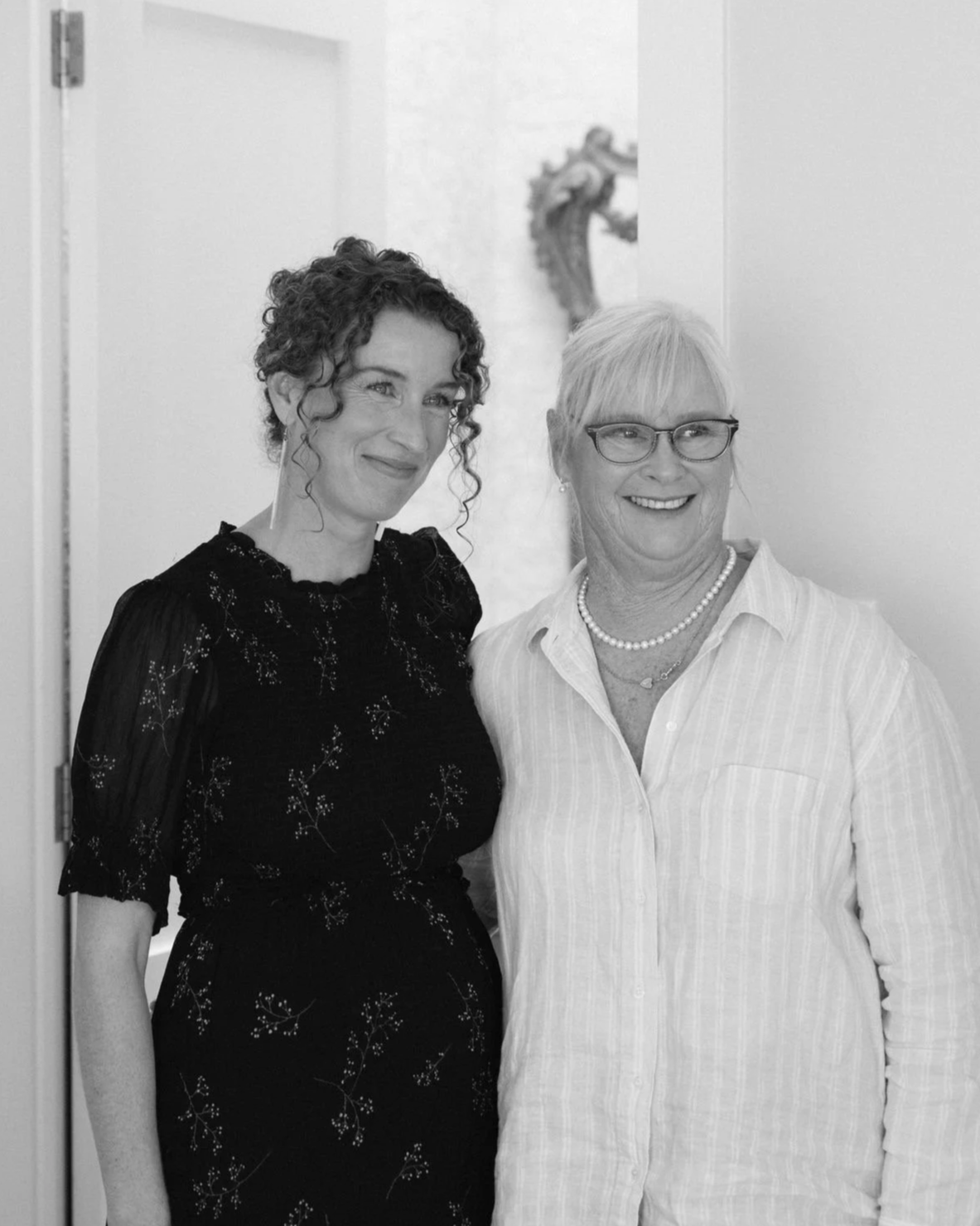 two women, one younger, one older, smile at something beyond the camera frame
