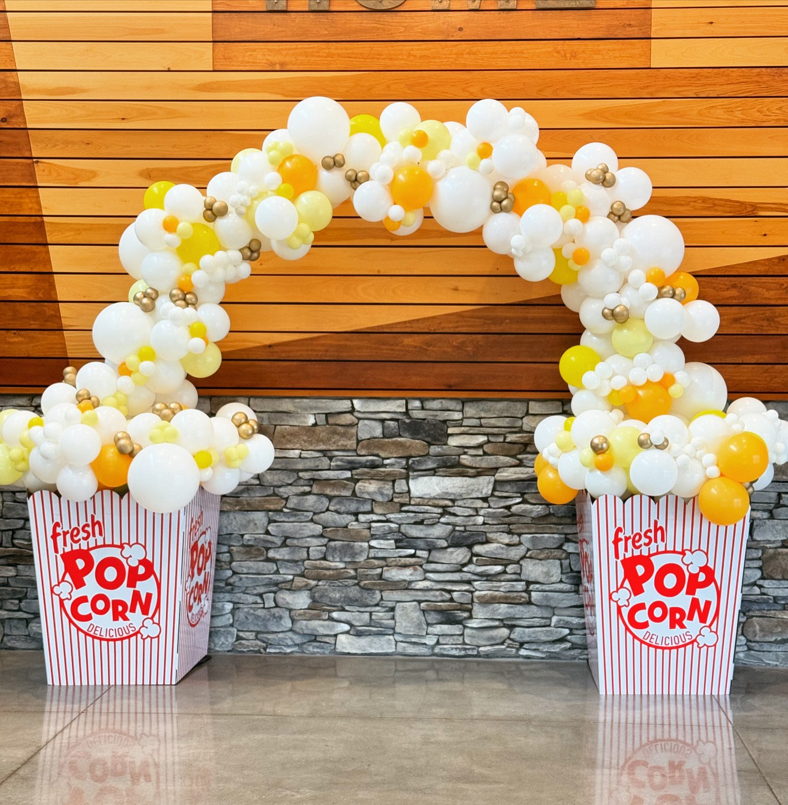 Balloon arch decorated with white, yellow, orange, and metallic gold balloons, supported by two boxes of popcorn with red and white stripes reading 'fresh popcorn delicious,' set against a wooden wall and stone accent wall.