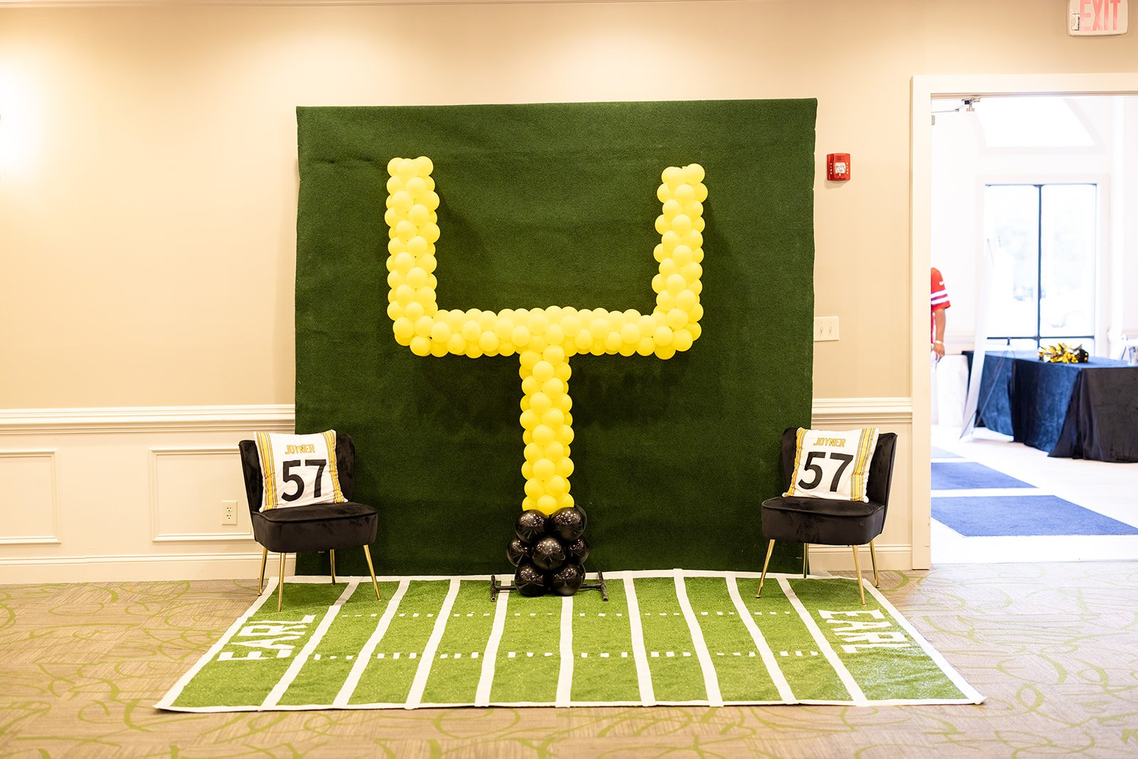 Decorative sports-themed display with a green grassy backdrop featuring a large yellow balloon number 4, two black chairs with sports pillows, and a green and white football field rug.