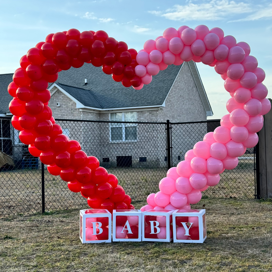 Heart-shaped balloon decoration with red and pink balloons and blocks spelling "BABY" at the base, set outdoors in a backyard.
