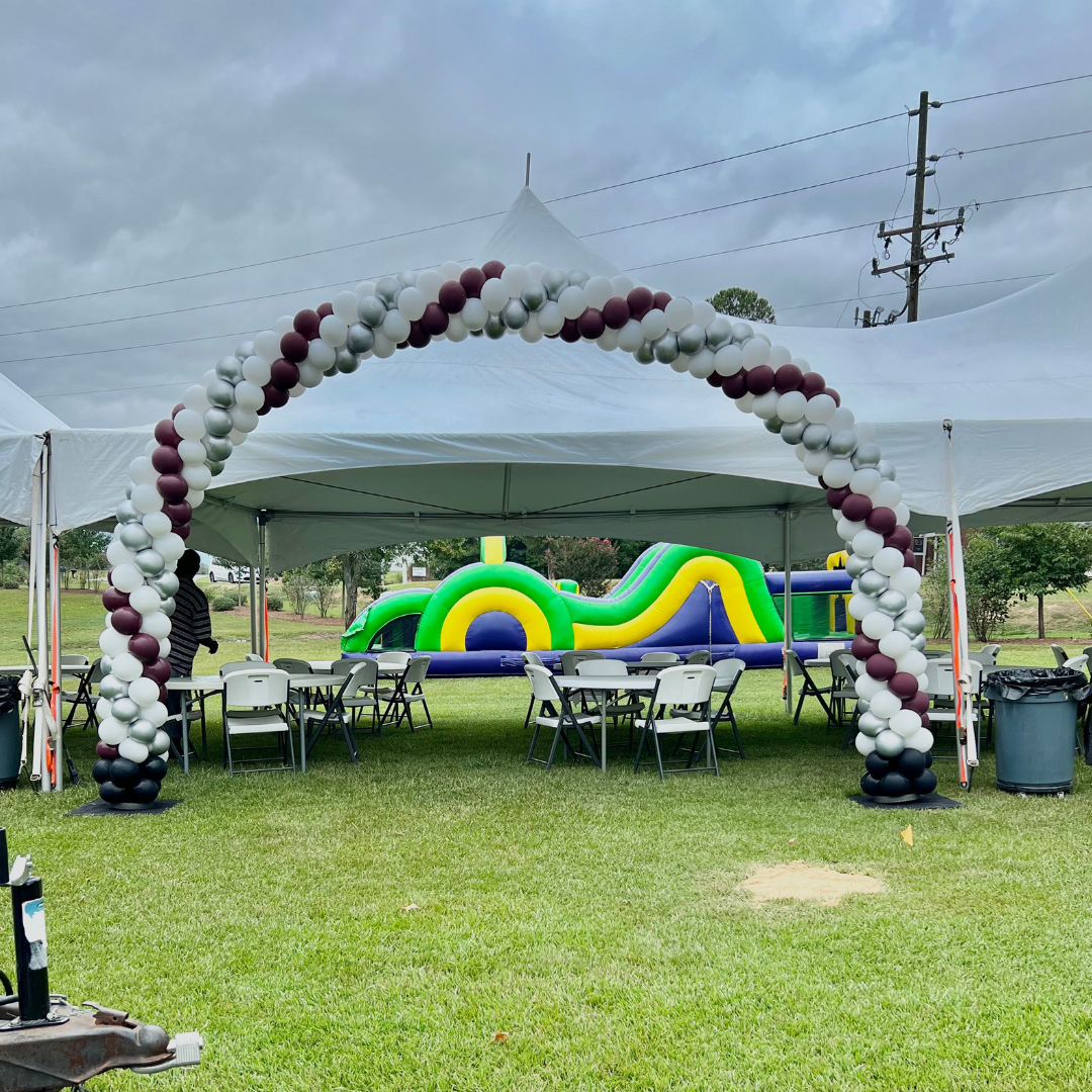 Outdoor decorated party area with white tents, a balloon arch in maroon, white, and silver, tables and chairs, and a colorful inflatable slide in green, yellow, and blue on a grassy field under a cloudy sky.