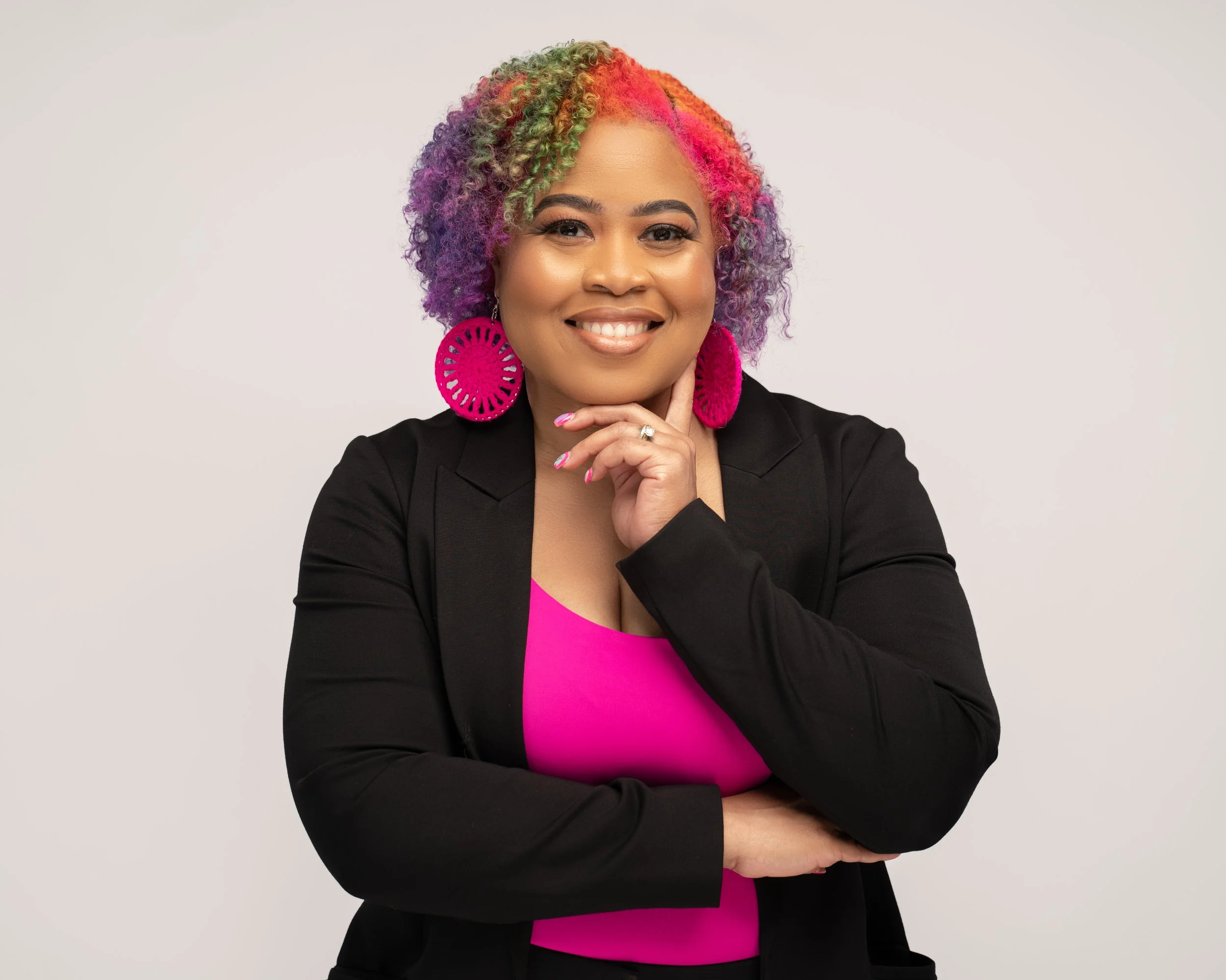 Woman with multicolored curly hair, pink top, black blazer, large pink earrings, smiling, posing with her hand on her chin.