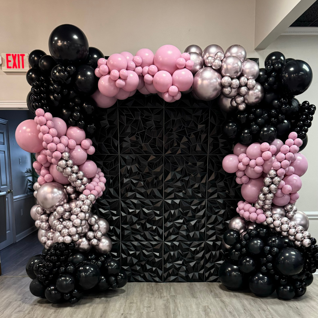 Balloon arch with black, pink, and silver balloons in front of a geometric black backdrop, near an exit sign in an indoor space.
