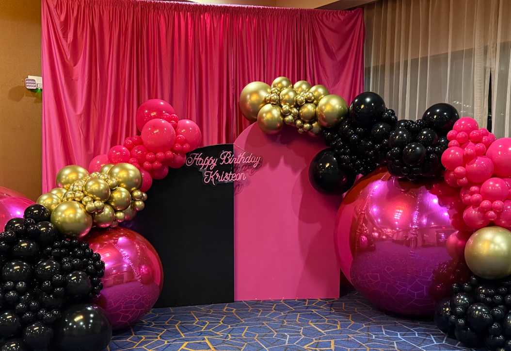 Decorative balloon arch with pink, black, and gold balloons around a pink backdrop with a blackboard that says 'Happy Birthday Kristaon', in a room with a patterned carpet and pink curtains.