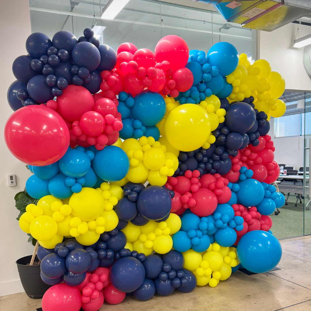 Colorful balloon display with pink, blue, yellow, and purple balloons of various sizes arranged in a cluster inside an office space.