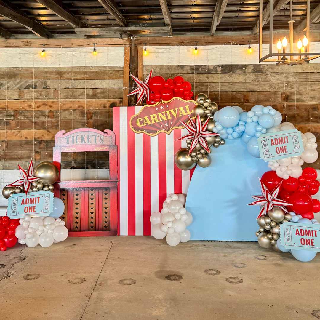 Decorative carnival-themed backdrop with red, white, blue, and gold balloons, a "Tickets" booth, a striped red and white panel with a "Carnival" sign, star-shaped balloons, and "Admit One" tickets on a rustic wooden wall background.
