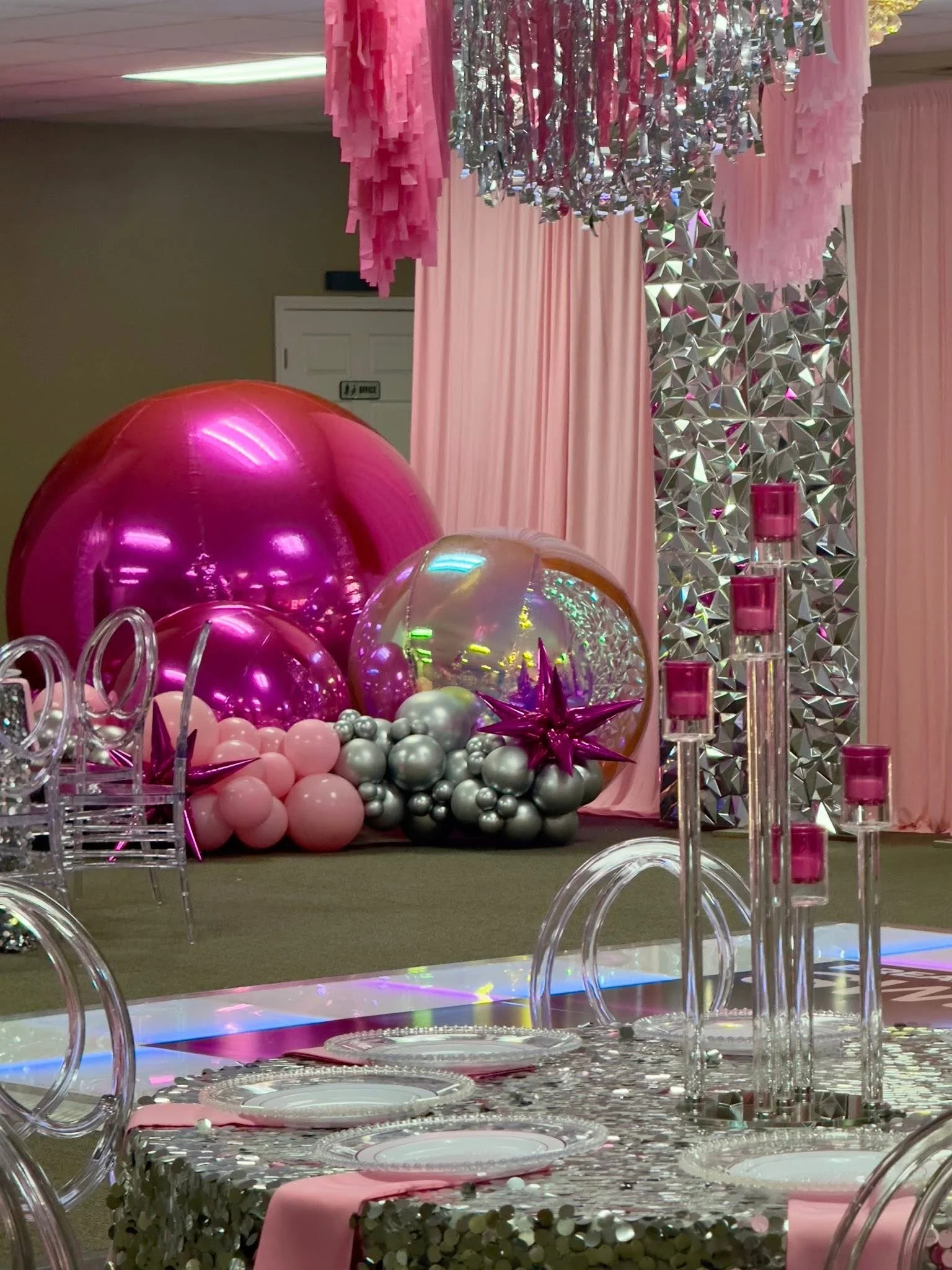 Party decorations with pink, silver, and iridescent balloons, a pink backdrop with curtain, hanging pink and silver streamers, and a table set with transparent chairs, pink napkins, and a sequin tablecloth.