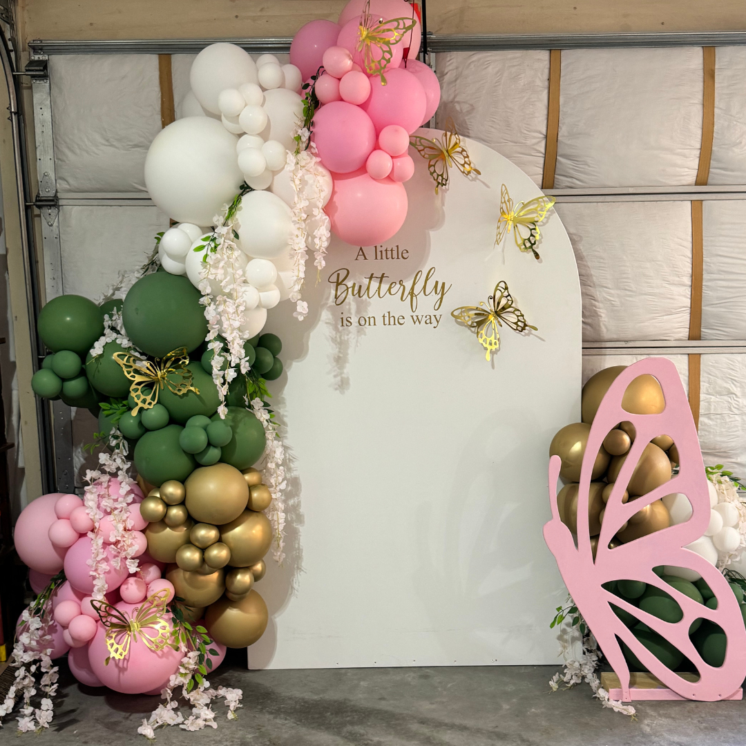 Decorative butterfly-themed balloon arch with pink, white, green, and gold balloons, floral accents, and paper butterflies around a white sign that reads 'A little Butterfly is on the way'.