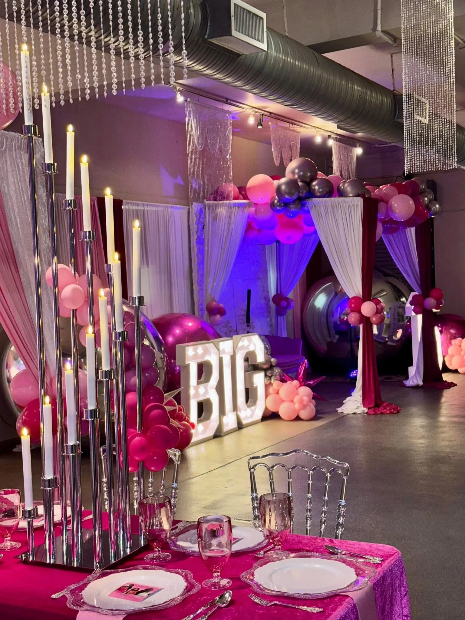 Decorated event space with pink, purple, and silver balloons, white drapery, illuminated marquee letters spelling 'BIG,' tall candlesticks with candles, and pink table settings with glasses and utensils.