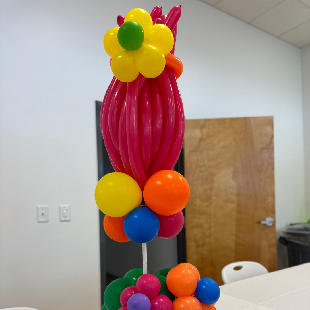 Colorful balloon bouquet with pink, yellow, orange, green, blue, and purple balloons arranged on a table in an indoor room.