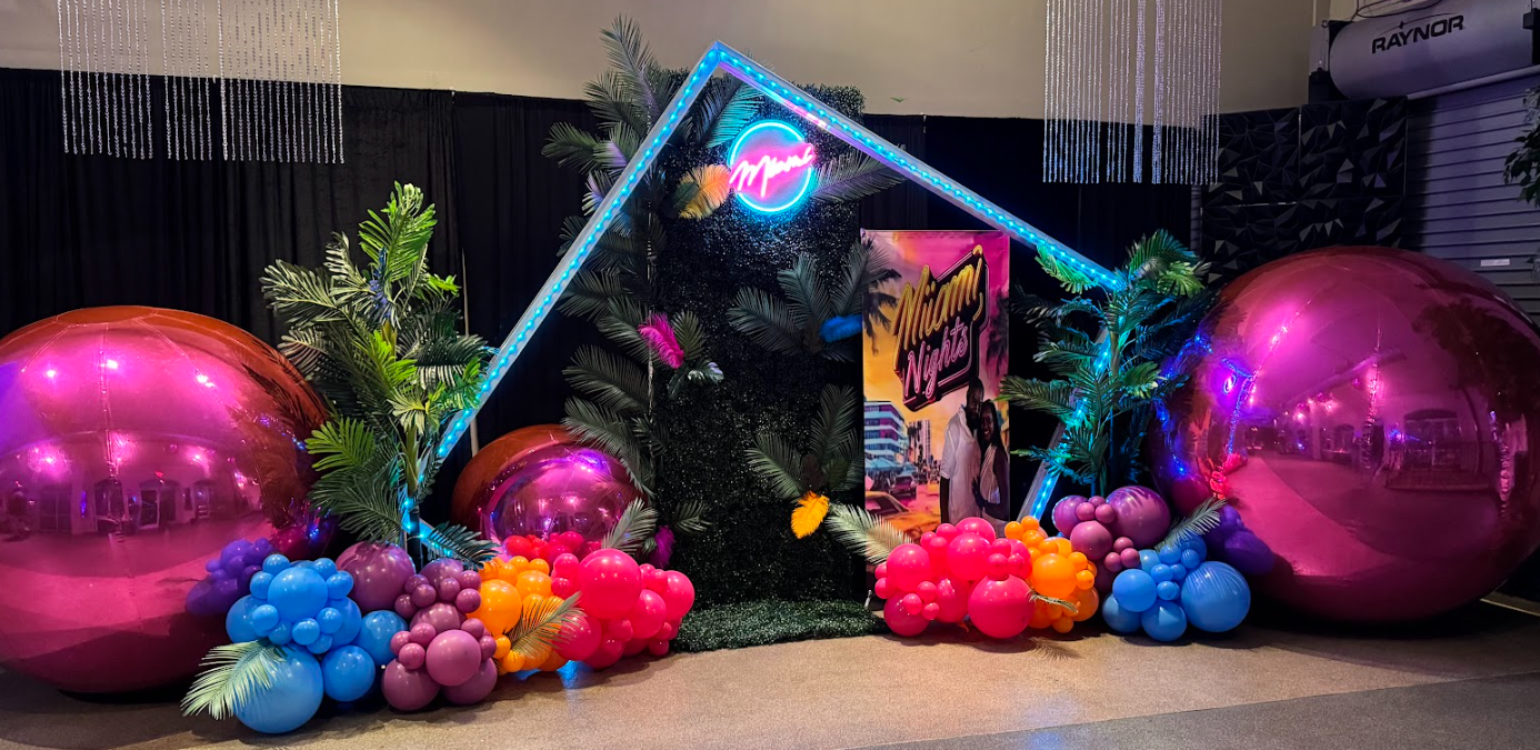 Decorative Miami Nights themed display with large pink metallic spheres, colorful balloons, tropical greenery, neon signs, and a promotional poster for Miami Nights.