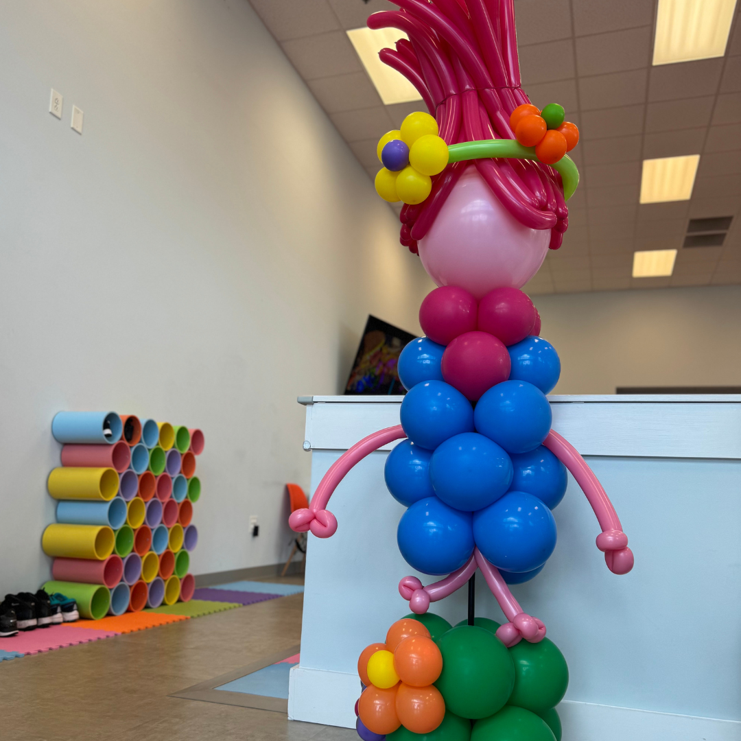 Colorful balloon sculpture of a woman with pink hair, adorned with flowers, placed in an indoor recreational or play area.