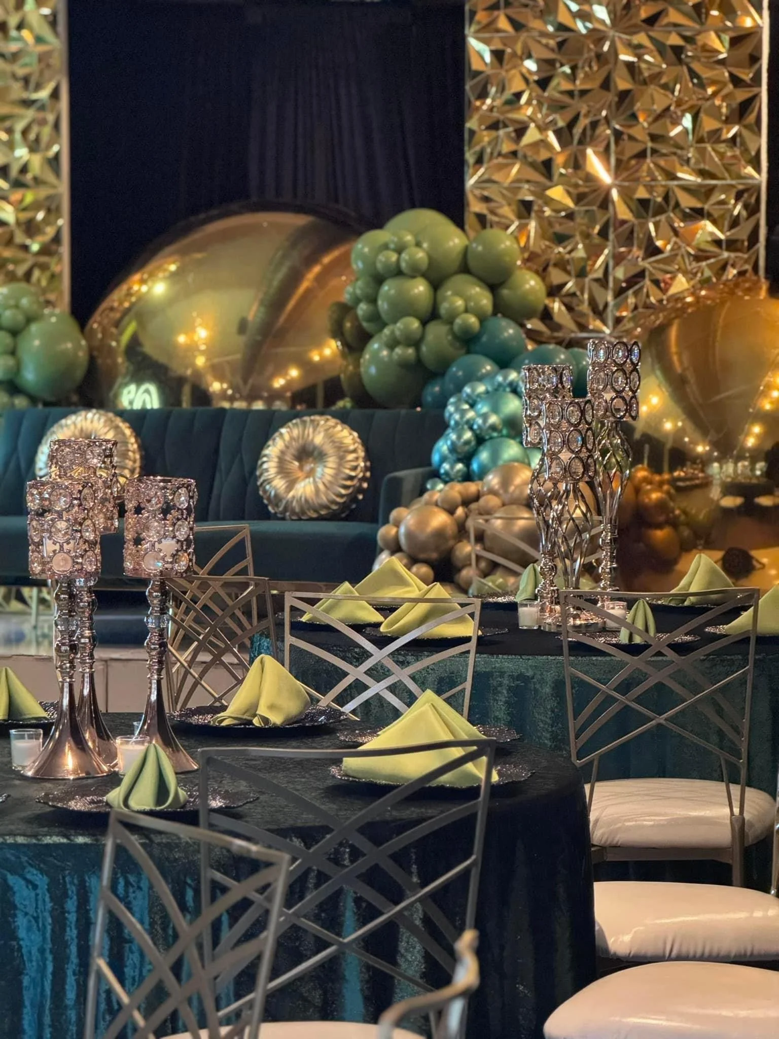 Elegant indoor event setup featuring a dark table with neon green napkins, ornate silver and crystal candle holders, and a backdrop decorated with gold, silver, and green balloons, reflective gold geometric panels, and plush navy velvet seating.