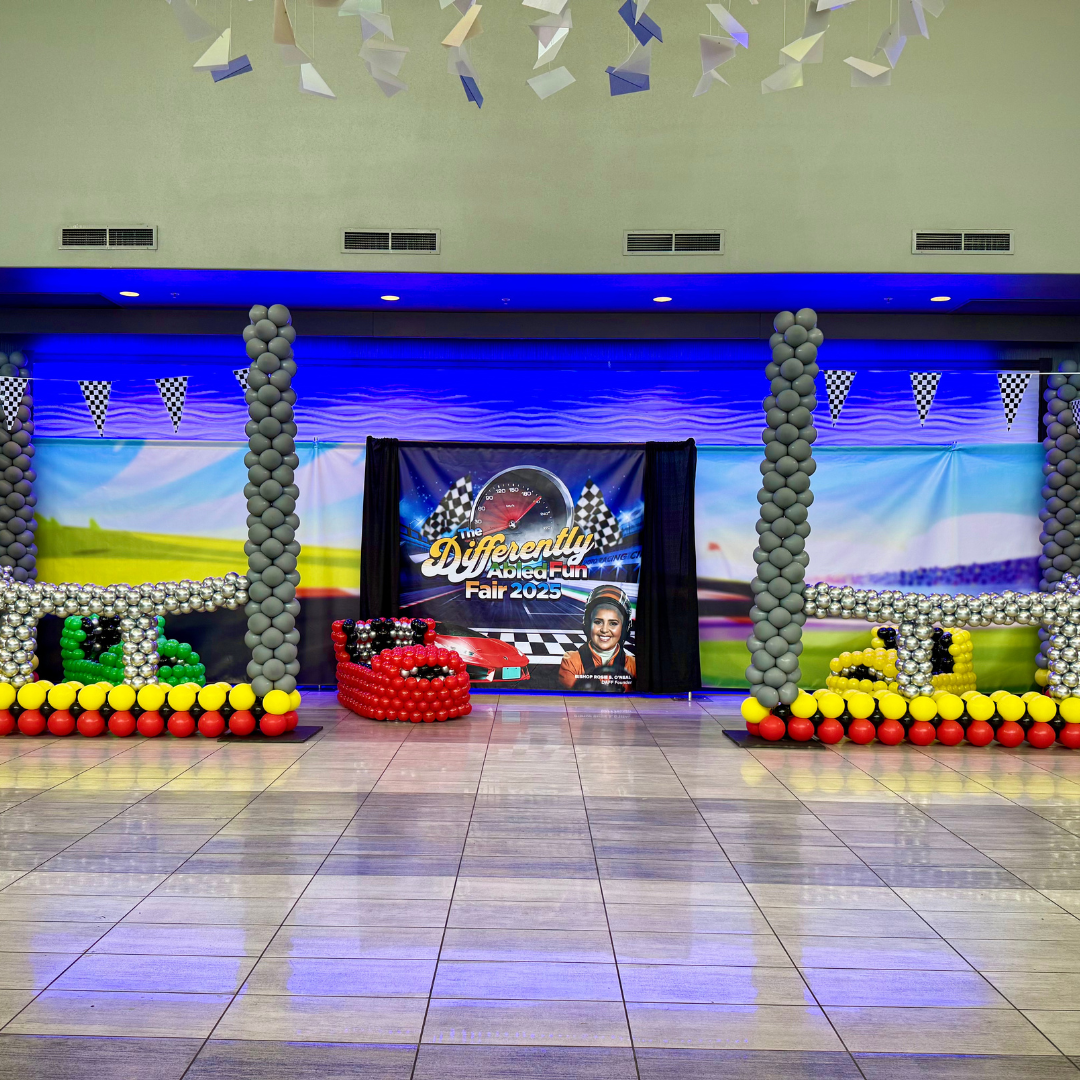 Balloon display with checkered flag banners, a backdrop with a racing theme, and a sign that reads 'The Differently Abled Fun Fair 2025' featuring a woman and a race car.