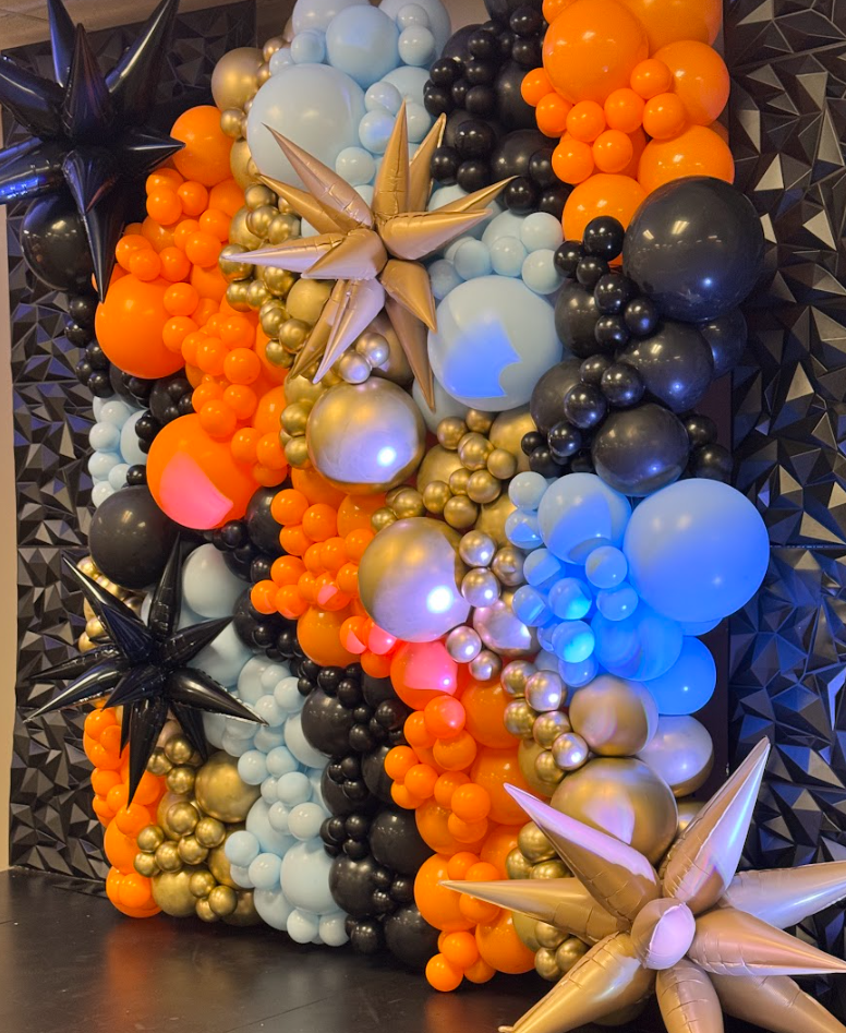 Colorful balloon arrangement with orange, black, gold, and light blue balloons, accented by star-shaped and cone-shaped balloons, against a geometric black wall.