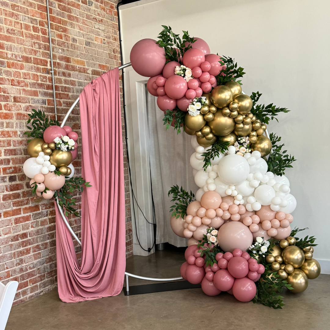 Decorated floral balloon arch with pink, white, gold, and nude balloons, accented with greenery and white flowers, with a pink fabric backdrop on a white circular stand.