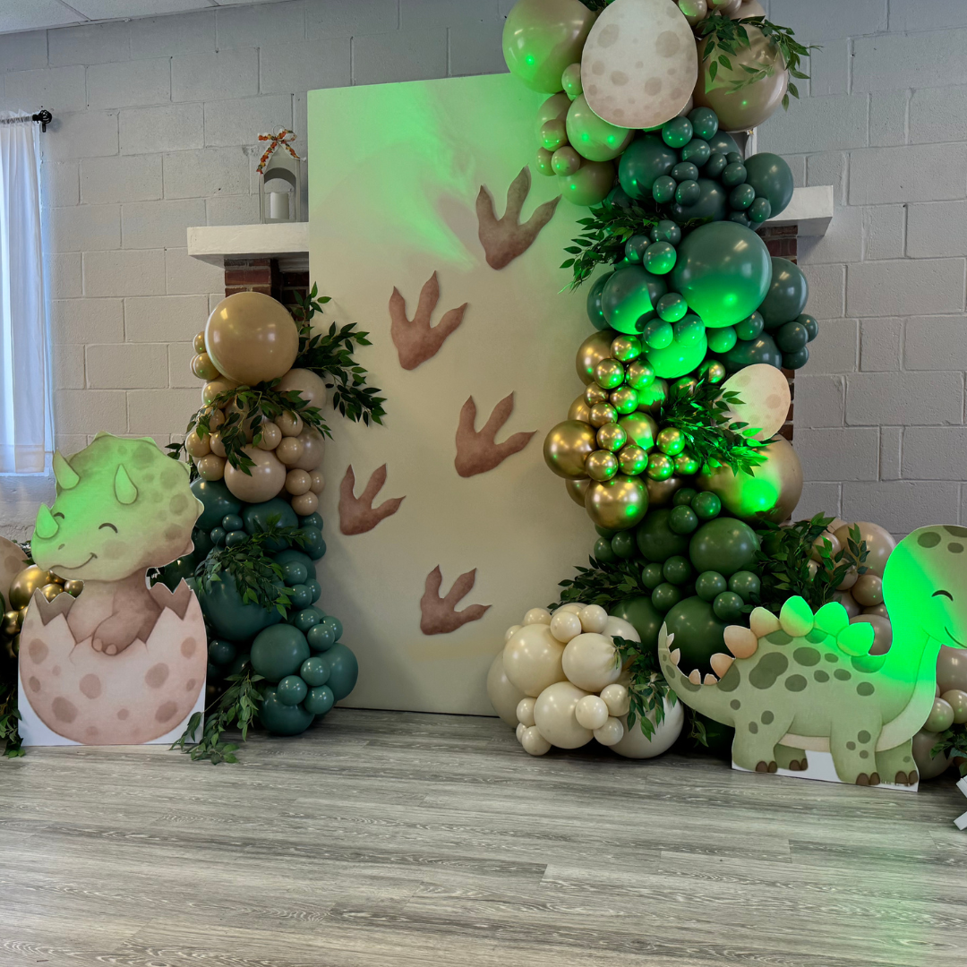 Decorative setup featuring green, white, and gold balloons arranged around a backdrop with cutout dinosaur and hatching dinosaur images. Green lighting illuminates the scene, which includes cartoon dinosaurs and playful dinosaur-themed elements.
