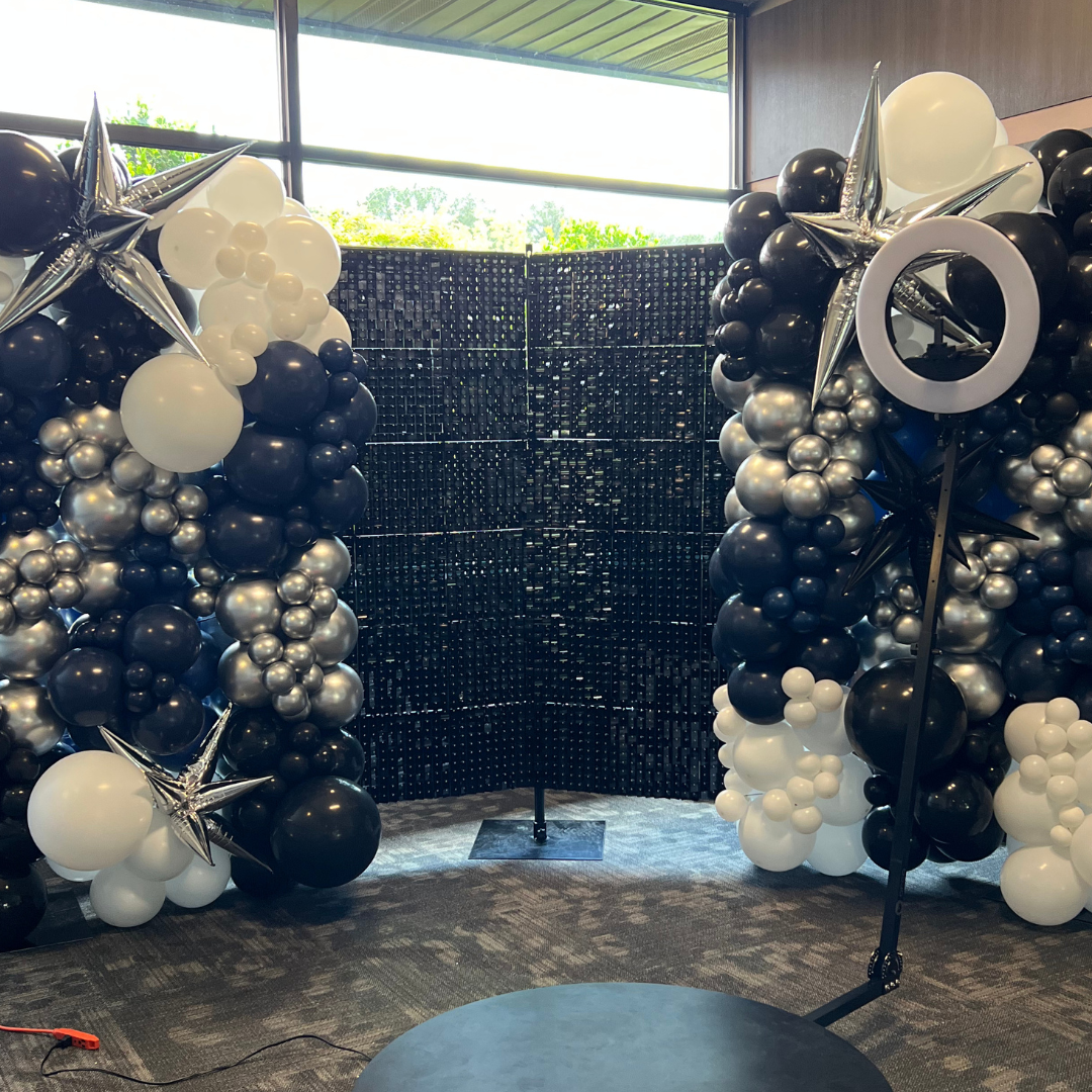 Decorative balloon arch with black, white, and silver balloons, accented with star-shaped balloons, set against a black backdrop with cheerful outdoor view through windows.