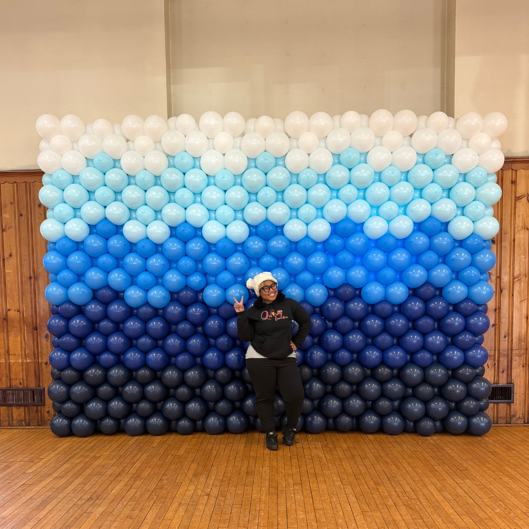 Woman standing in front of a large balloon backdrop arranged in horizontal color stripes from white, light blue, medium blue, to dark blue, indoors on a wooden floor.