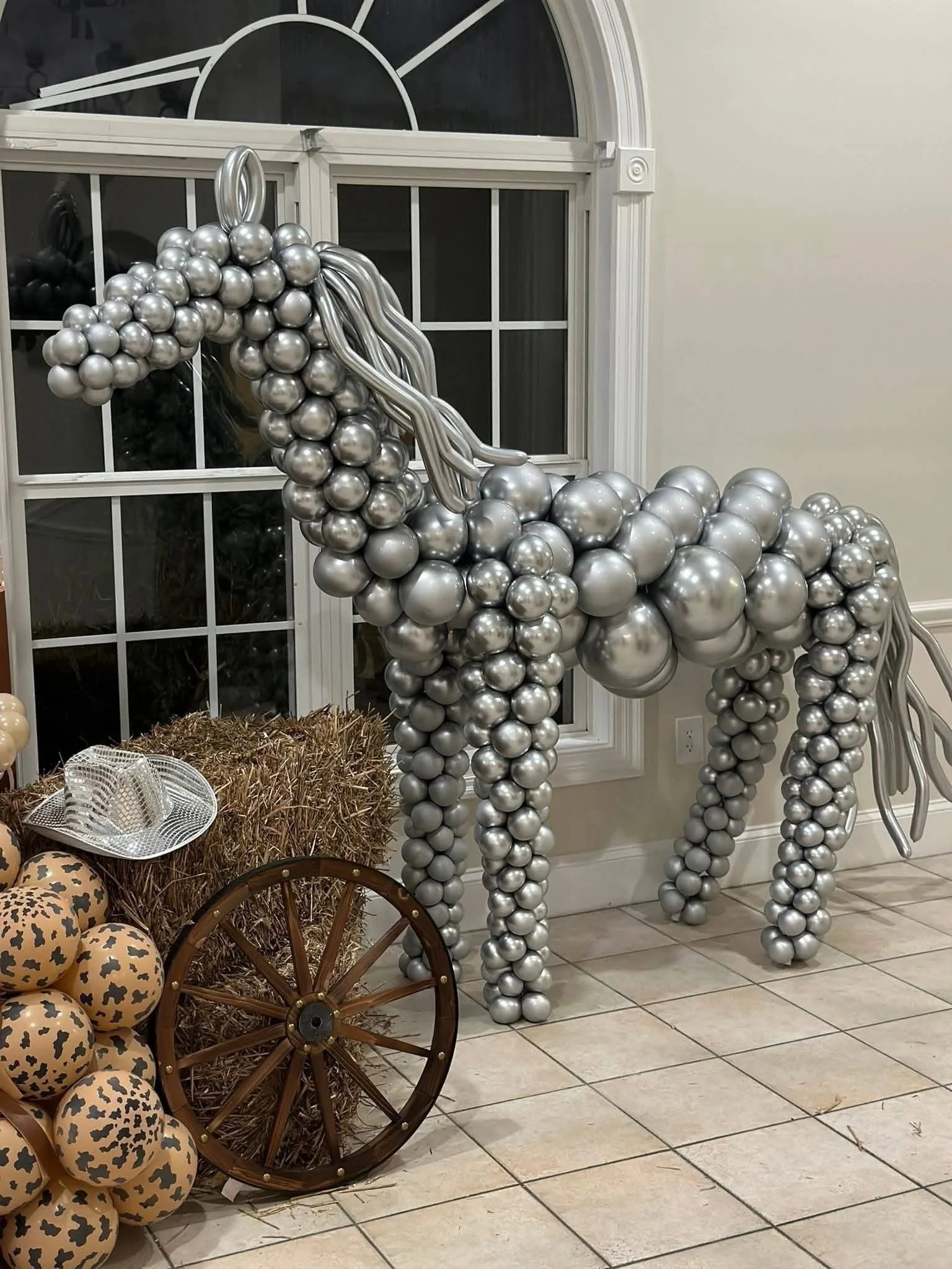 Decorative silver balloon sculpture of a horse, situated indoors near a window and a hay bale with soccer ball-patterned balls and a wagon wheel nearby.