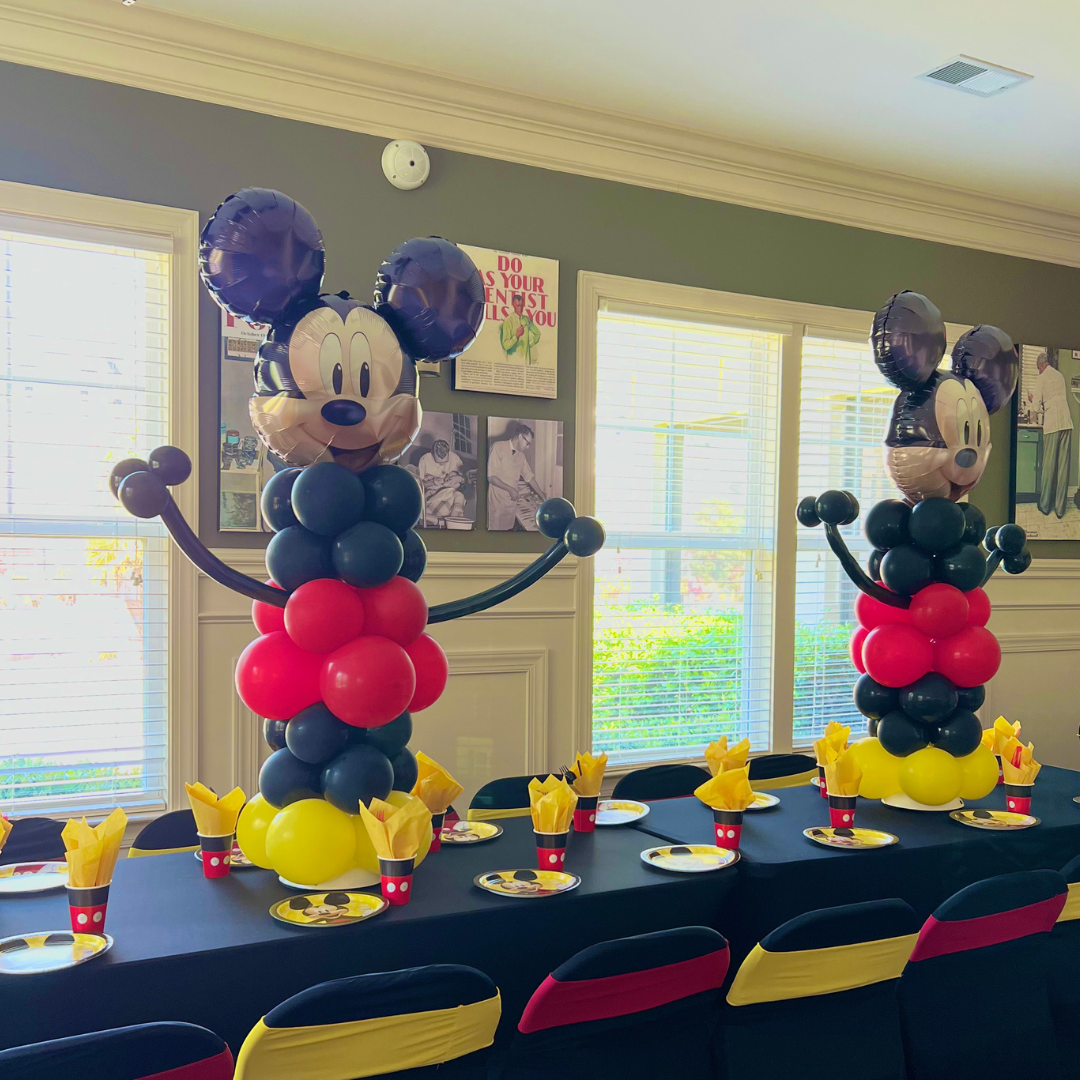 Party table setup with two large Mickey Mouse balloon sculptures as centerpieces, surrounded by themed plates and yellow napkins, in a room with windows and framed photos on the wall.