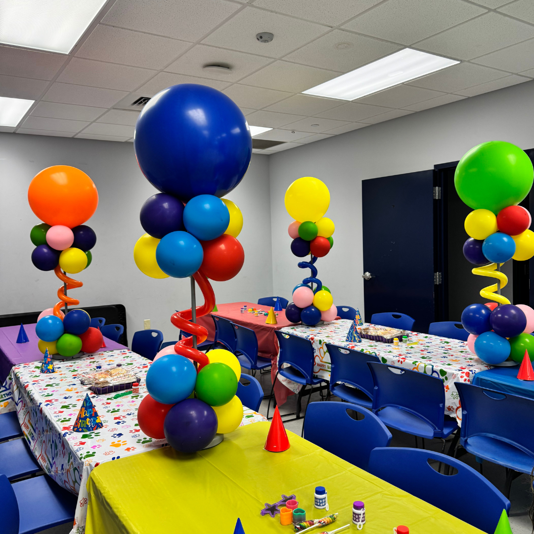 Colorful balloons arranged on tables for a birthday party with festive tablecloths, party hats, and party favors in a decorated event room.