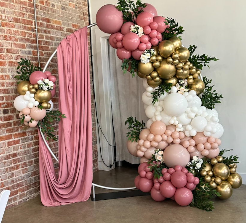 Balloon arch with pink, white, and gold balloons, decorated with greenery and white flowers, against a brick wall and pink curtain.