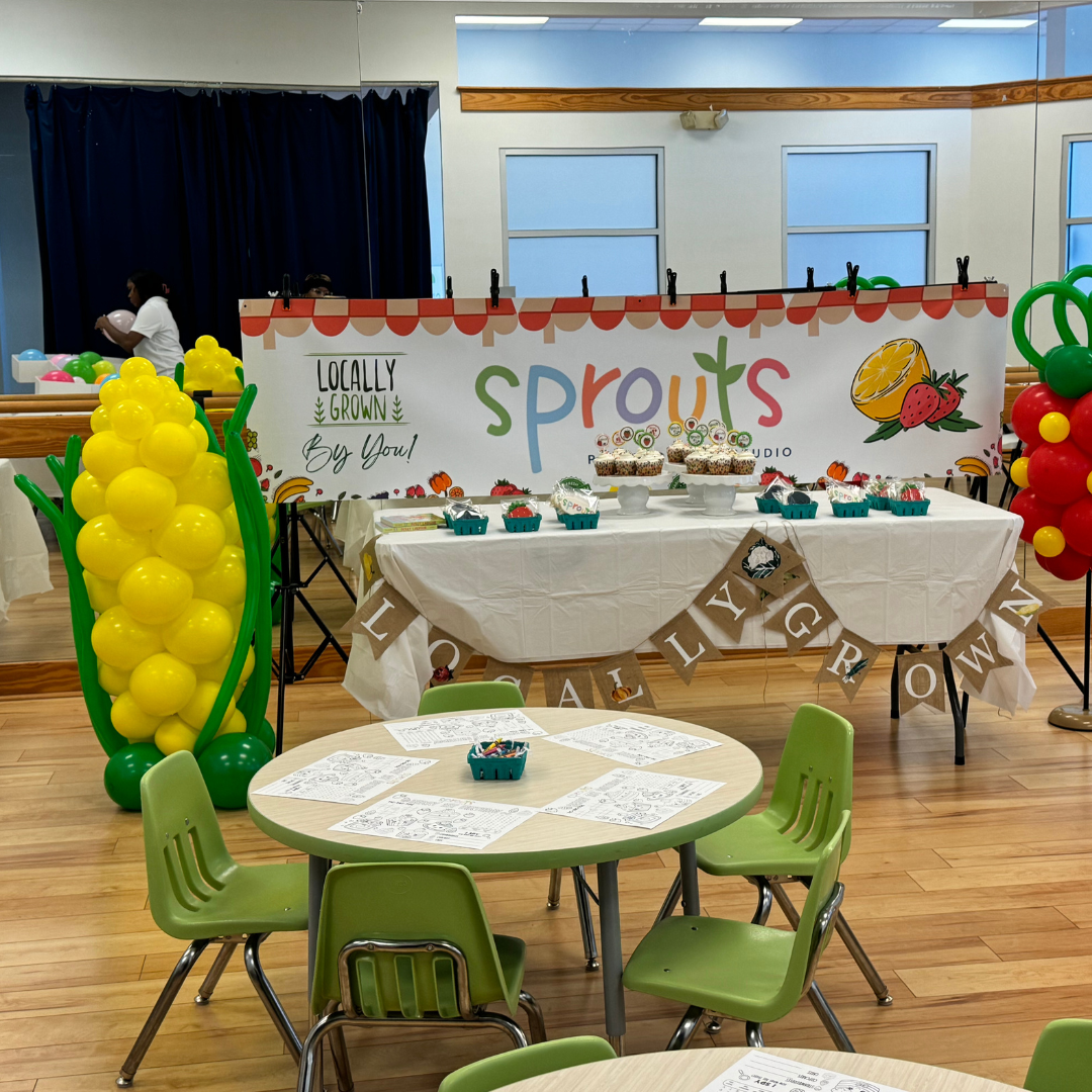Decorative table with cupcakes and treats, with a colorful banner reading 'Locally Grown' and 'Sprouts' behind it. Balloon arrangements on each side and a small round table with coloring sheets and crayons in the foreground inside a room with wooden floors and large windows.