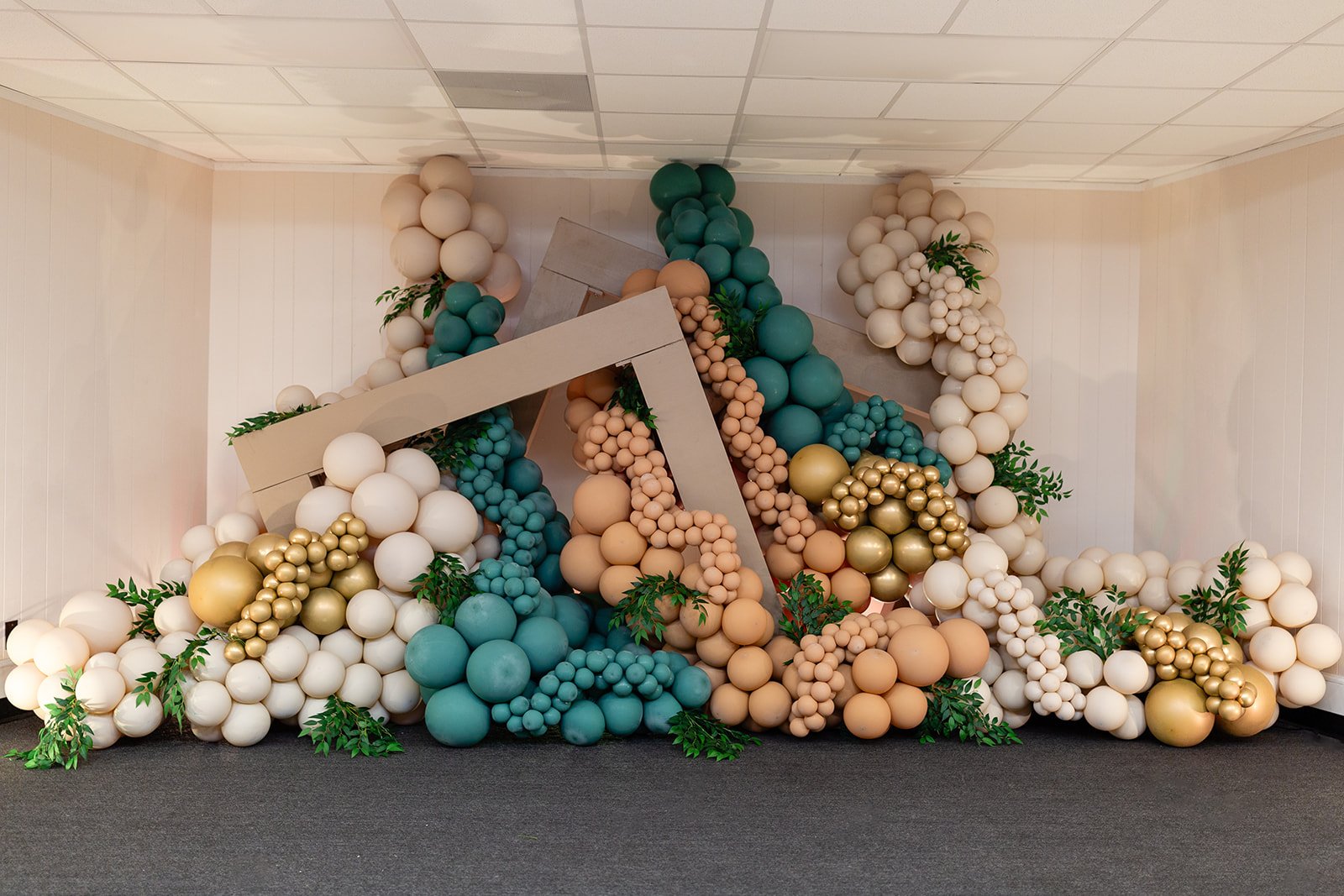 Decorative balloon display in shades of white, peach, teal, and gold, with green foliage, arranged in a cascading formation in an indoor space.