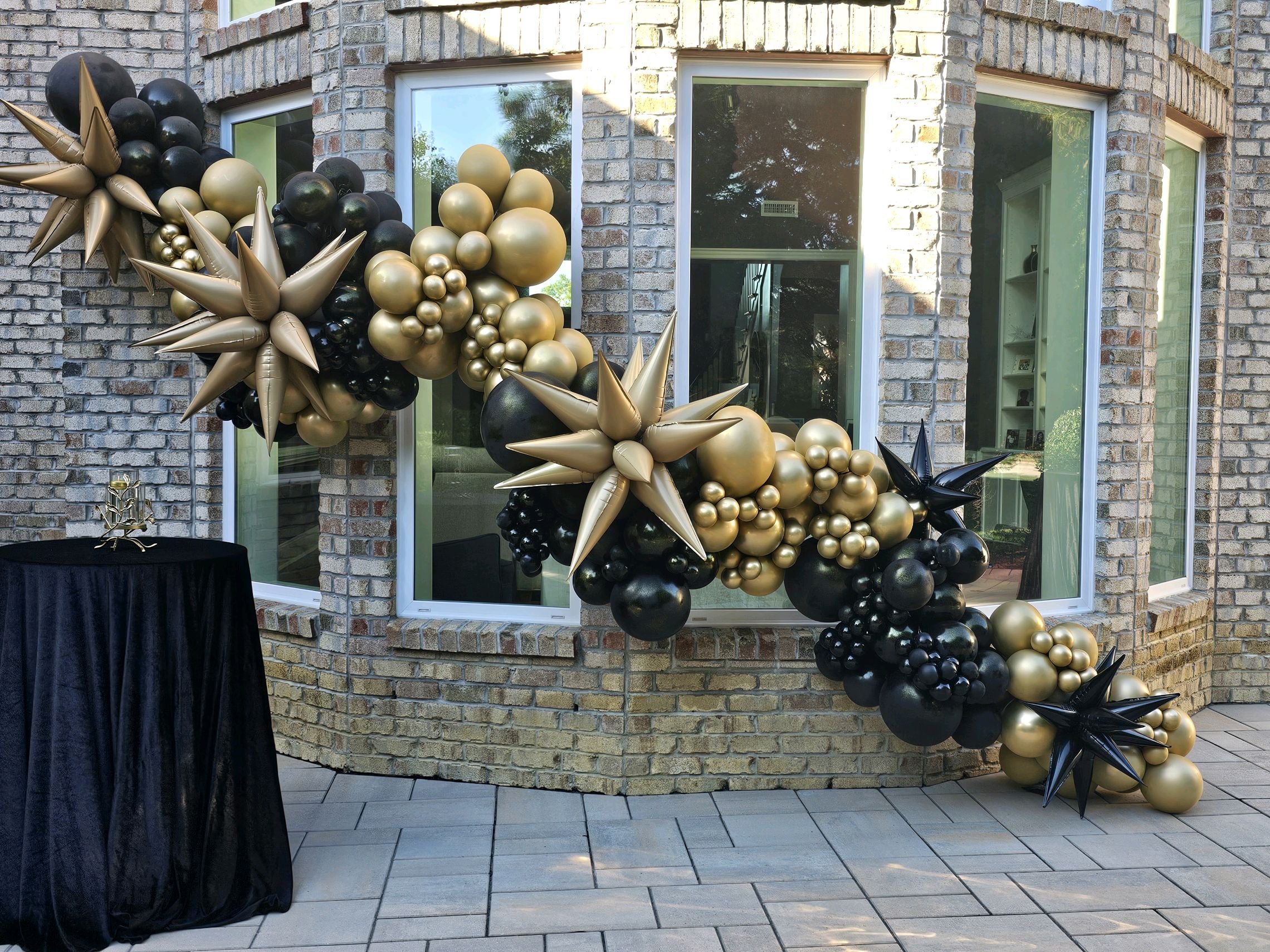 Black and gold balloon decoration arch on brick house exterior window wall, with a small black table and gold candle holder in front.