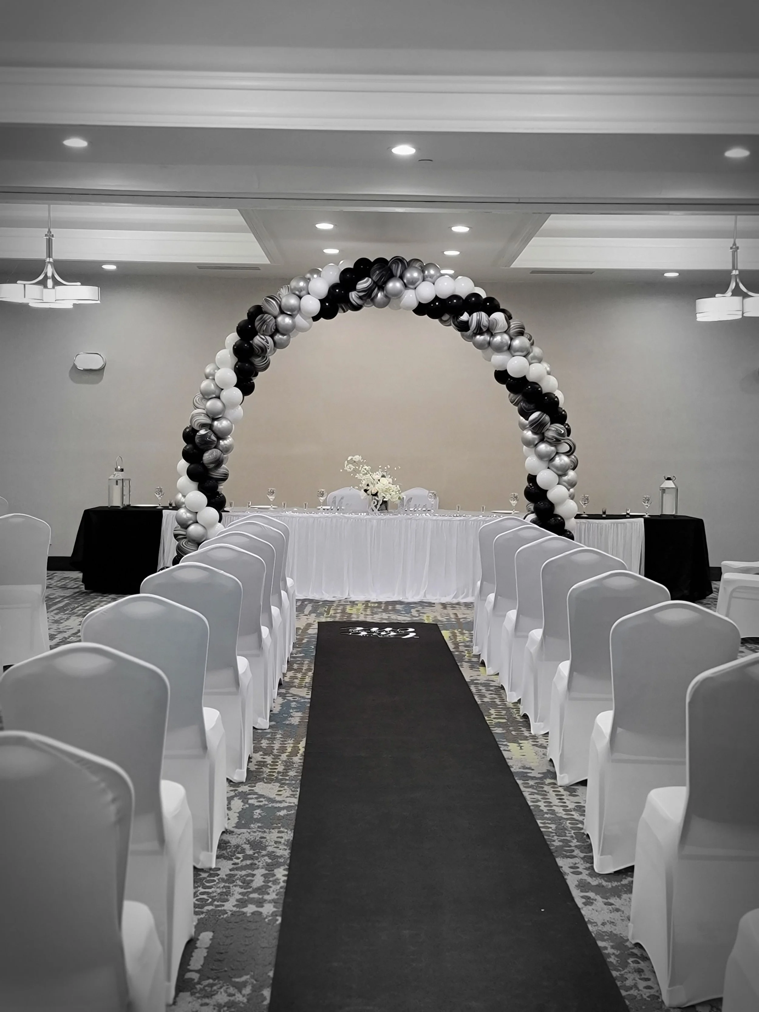 An indoor event space decorated with white chairs and a black aisle runner, with a balloon arch in black, silver, and white framing a decorated table at the front, set for a wedding or celebration.