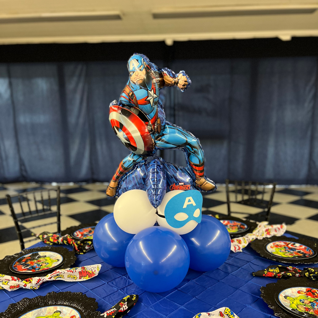 A superhero-themed birthday party table with a Captain America balloon centerpiece, featuring a foil balloon of Captain America in a punching pose, surrounded by blue, white, and a white skull-shaped balloon. The table is decorated with themed plates