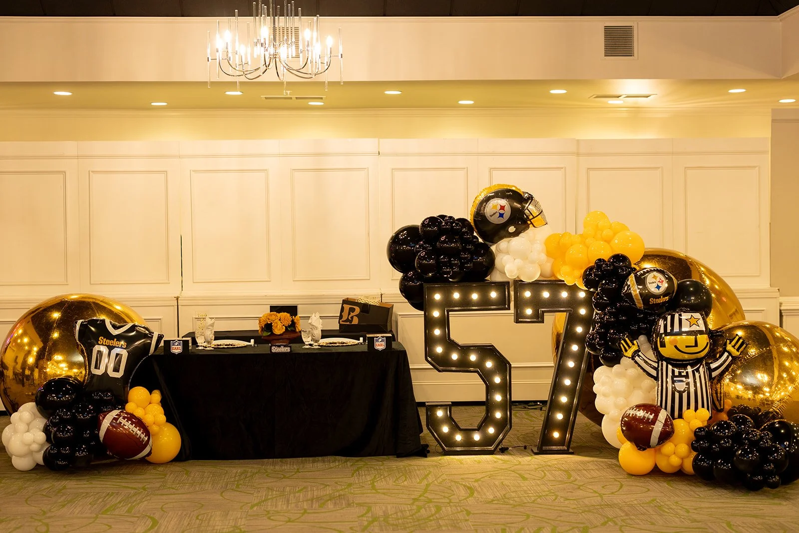 Decorative table setup for a Pittsburgh Steelers-themed celebration featuring large black and gold balloons, a football balloon, and lighted '57' numeral display, with a table holding floral arrangements and place settings in a banquet hall.