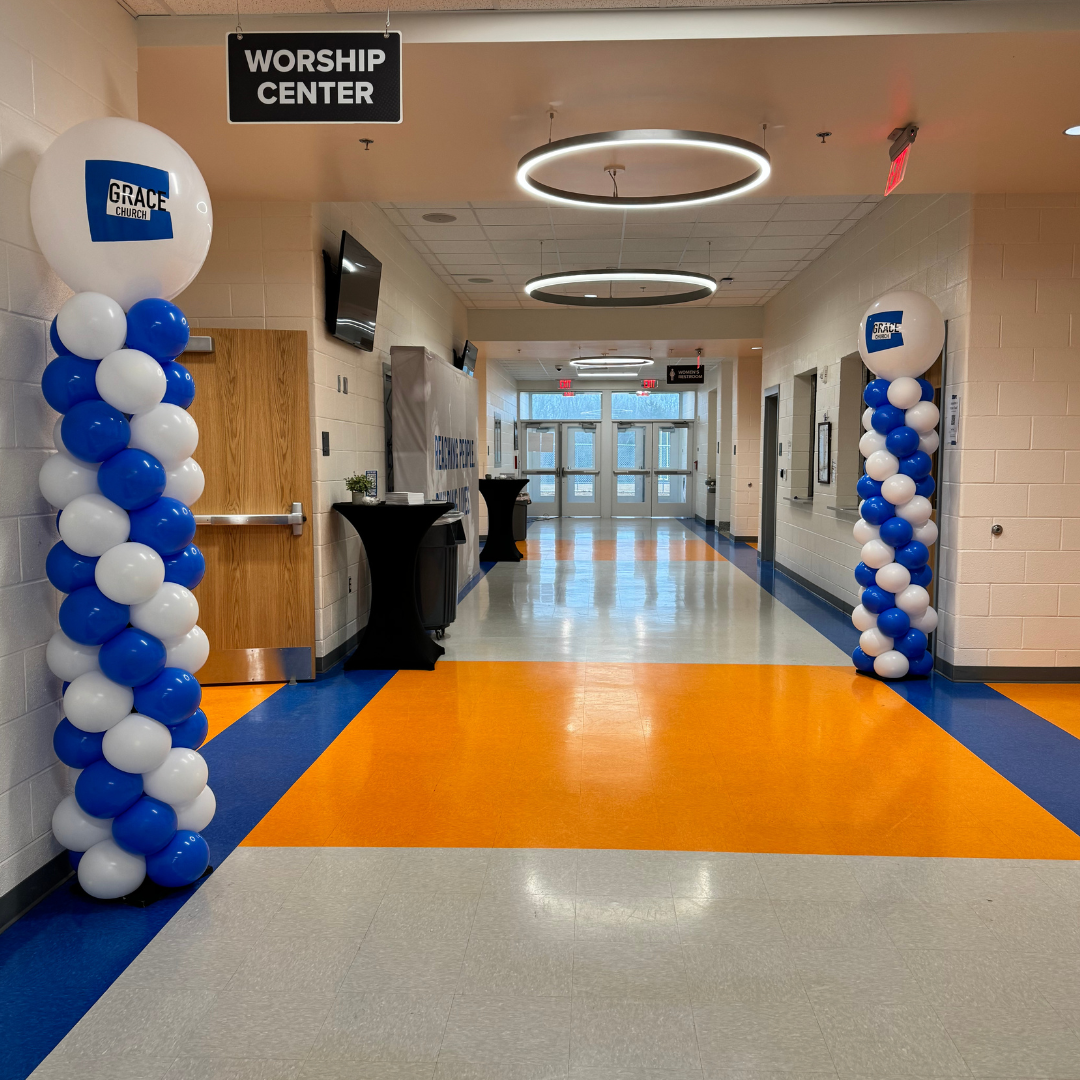 Interior of a church or community center lobby with a sign indicating the worship center, decorated with blue and white balloons on pillars, and a hallway leading to glass doors at the back.