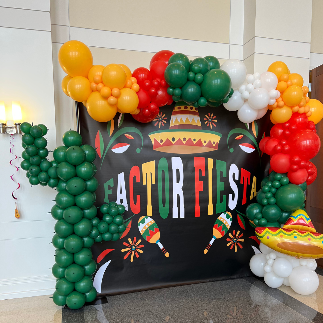 Colorful Fiesta-themed balloon arch with green, yellow, red, white, and multicolored balloons surrounding a Fiesta decorations backdrop featuring a sombrero, maracas, and festive patterns.