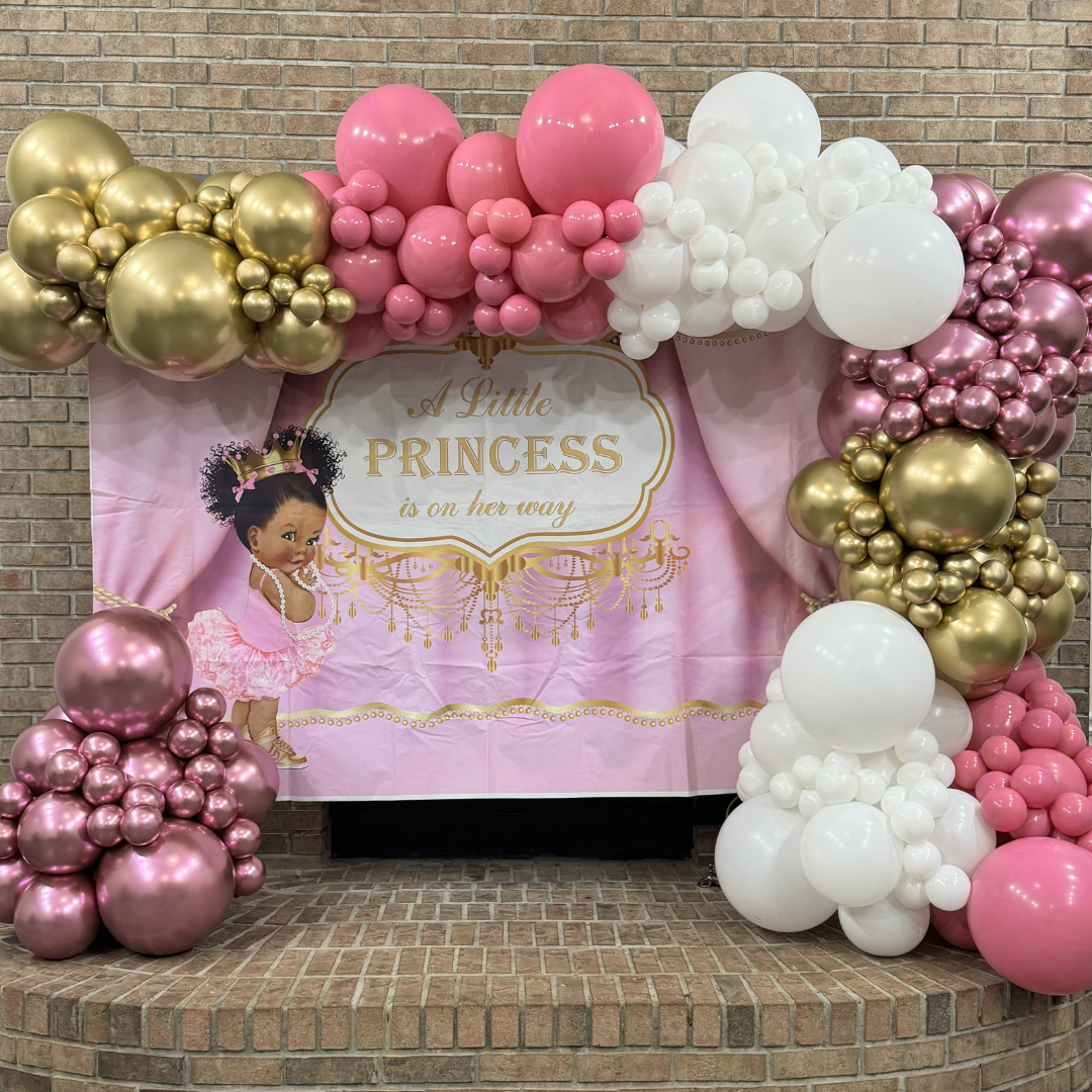 Decorative balloon arch with pink, white, and gold balloons surrounding a pink birthday backdrop with a princess theme, featuring a cartoon girl and the text 'A Little PRINCESS is on her way'.