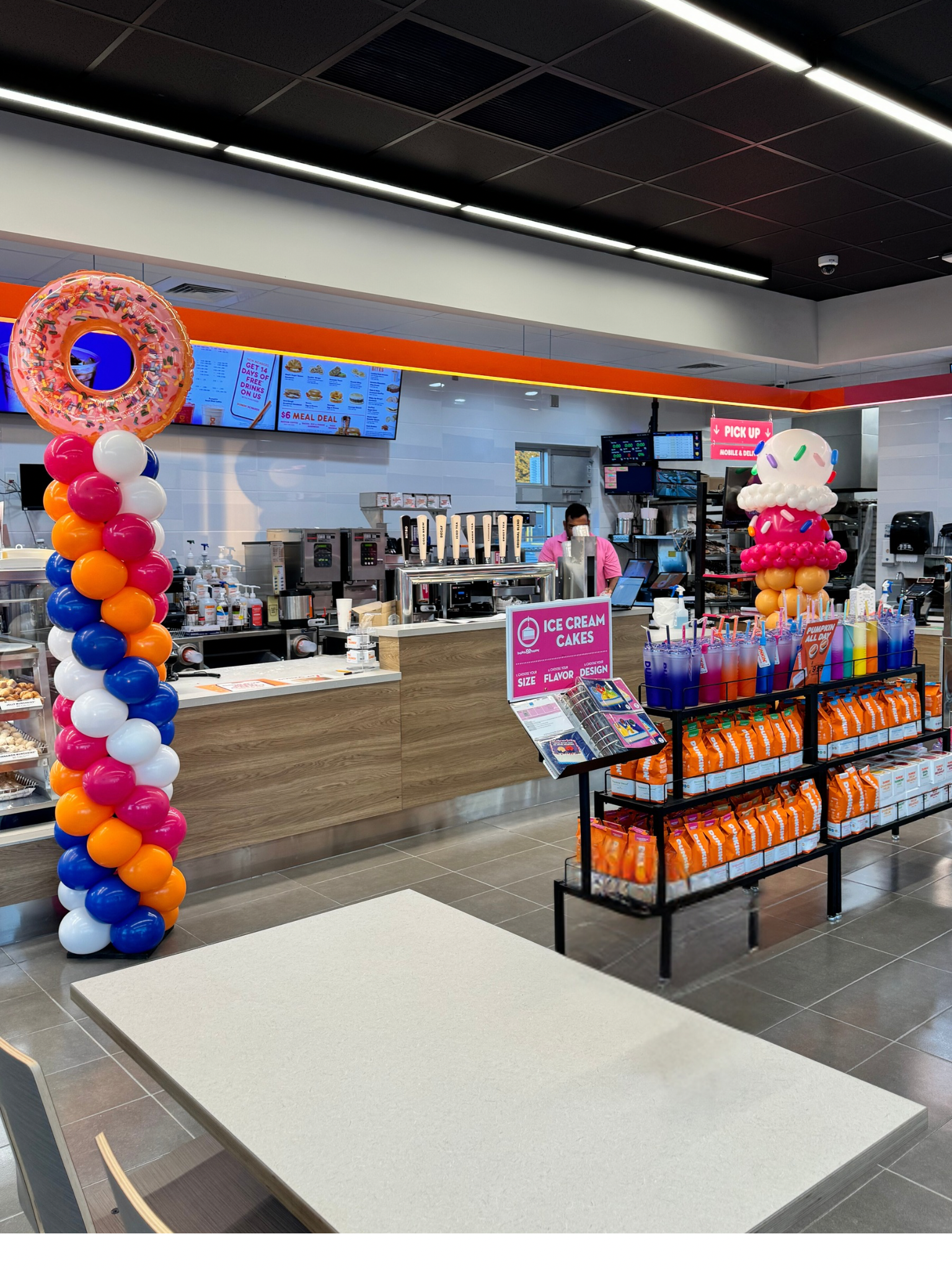 Inside a fast food restaurant decorated for a celebration, with colorful balloon arches themed like donuts and ice cream cones, and a display of syrups and snacks near the counter.
