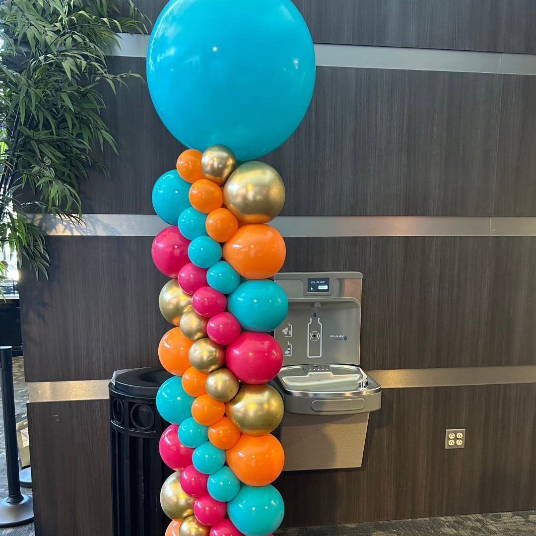 Colorful balloon column with blue, pink, orange, and gold balloons next to a water fountain and a trash can against a dark wood-paneled wall.